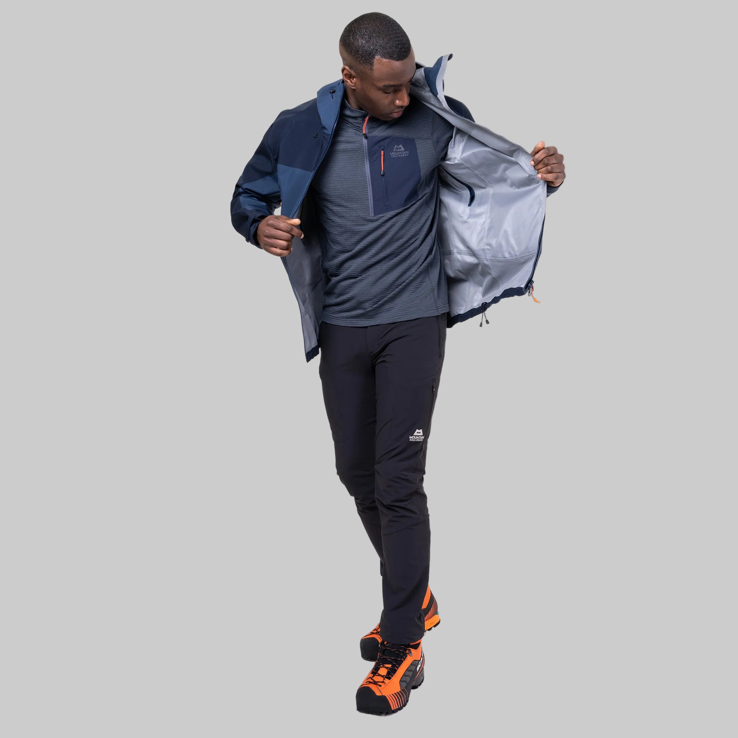 A man modeling a dark navy and gray technical jacket, shown opening the collar to reveal a light gray interior lining. He is also wearing dark gray athletic pants and bright orange and black trail running shoes, set against a plain white background.