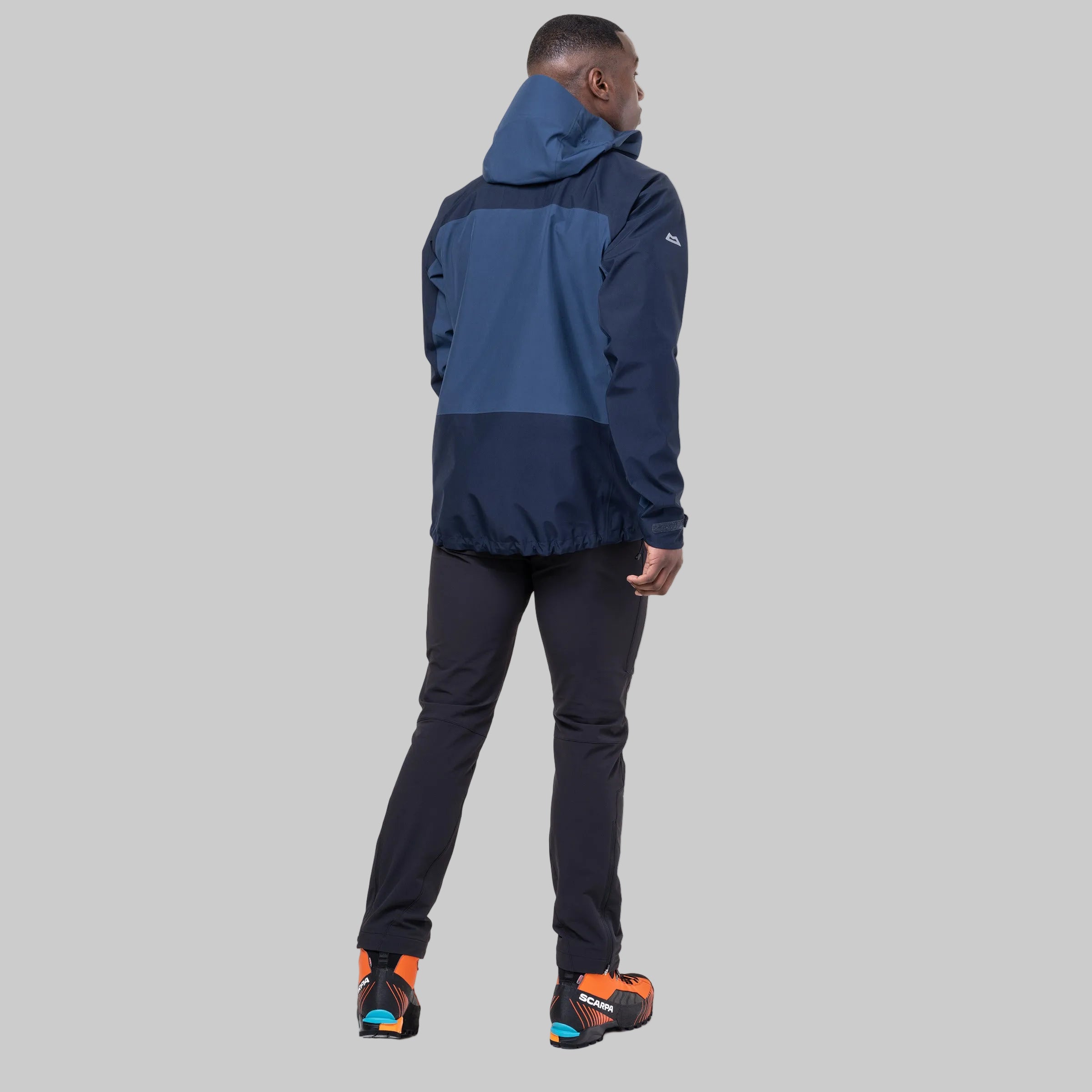 A man wearing a colorblock hooded jacket in navy and slate blue, paired with dark pants and orange and blue shoes, shown from a rear three-quarter view on a white background.