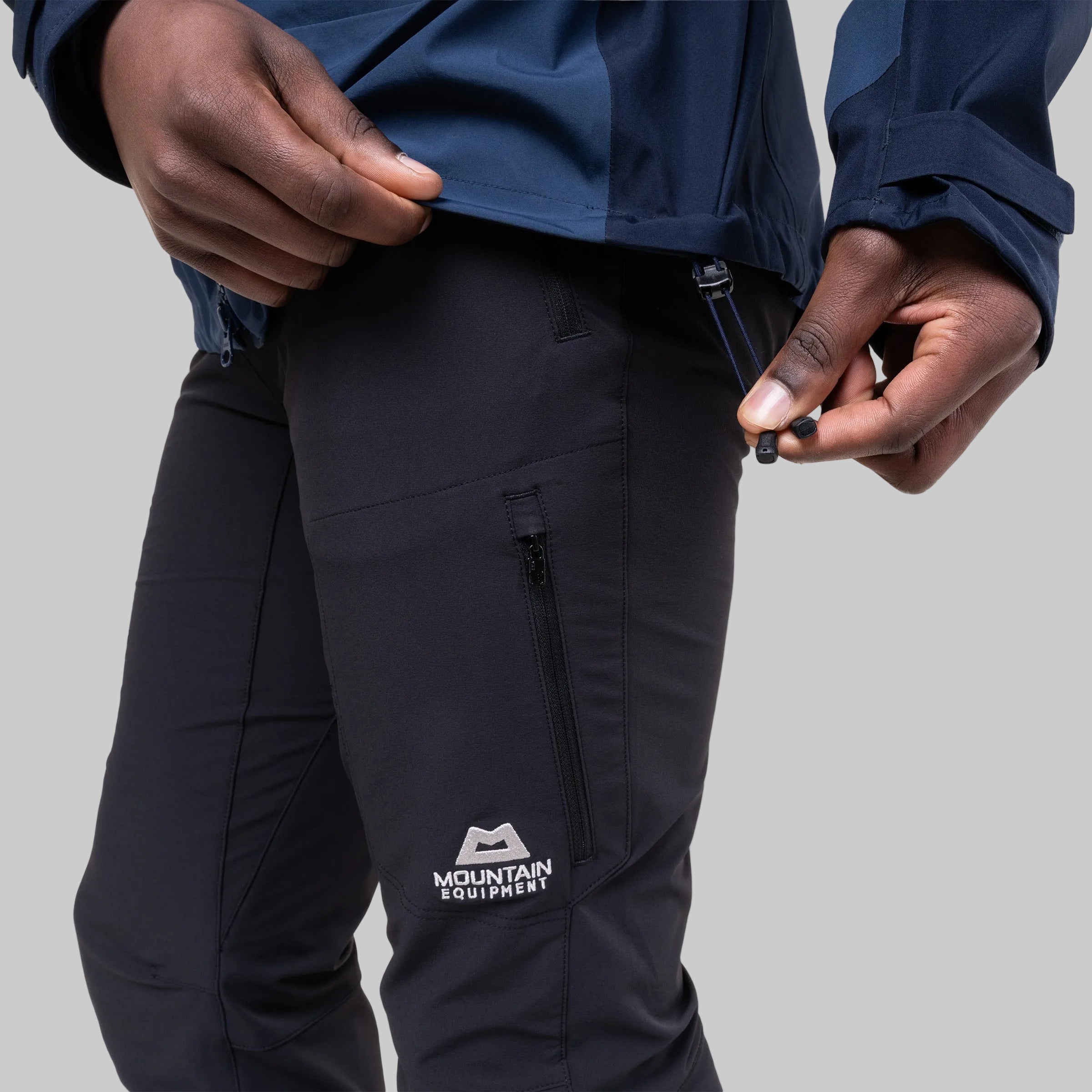 Close-up of a person wearing dark navy technical pants with a zippered pocket and a white Mountain Equipment logo, paired with a navy jacket against a white background.