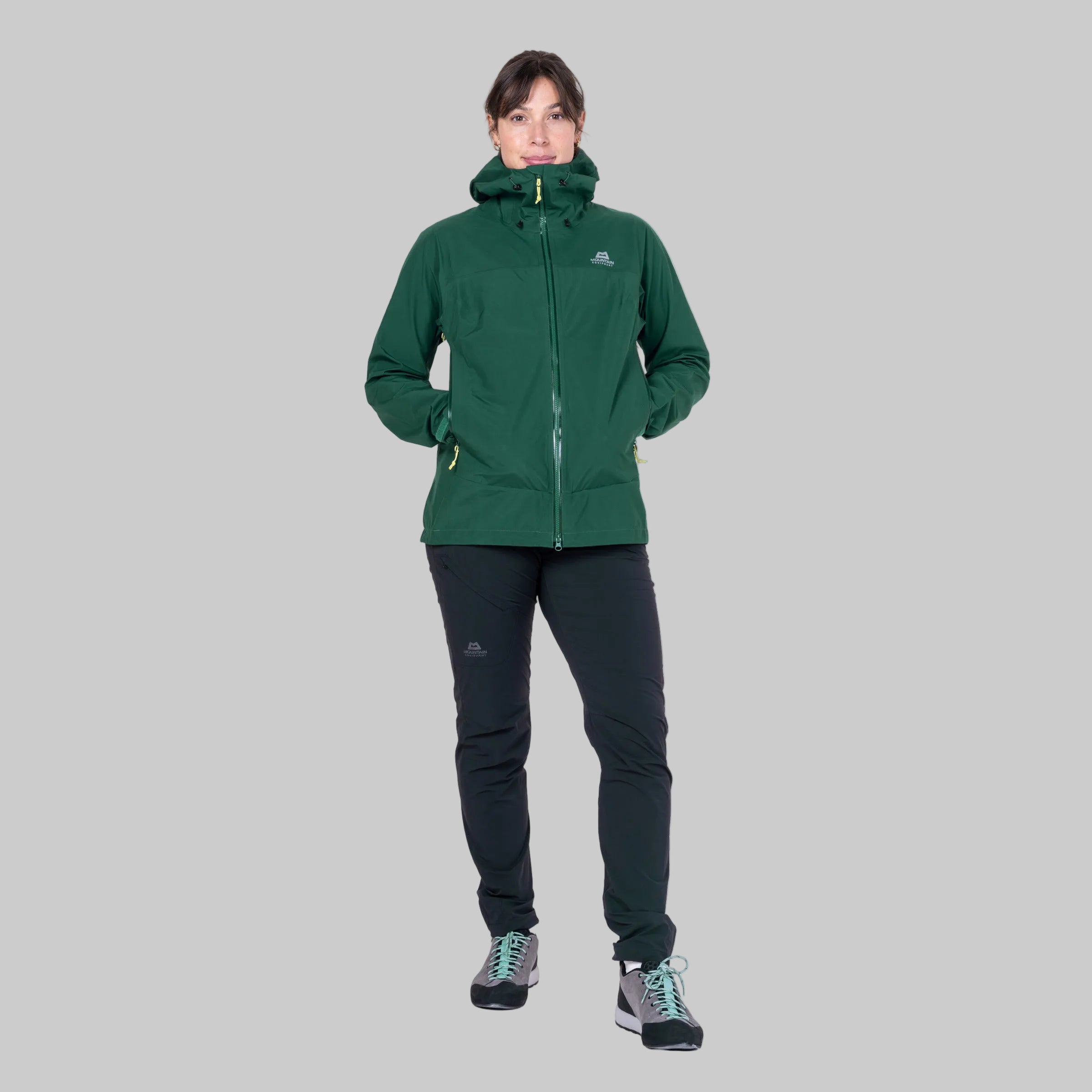 A woman modeling a dark green hooded technical jacket and black pants against a plain white background.