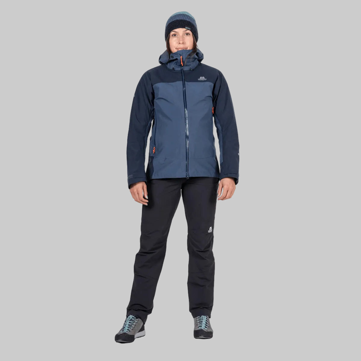 A model wearing a two-tone dark blue and navy hooded technical jacket, black pants, and a dark beanie, standing against a solid white background.