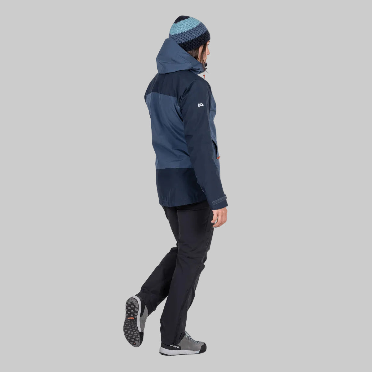 A man wearing a navy and blue hooded jacket, dark pants, and a blue and gray beanie, shown from the side against a plain white background.