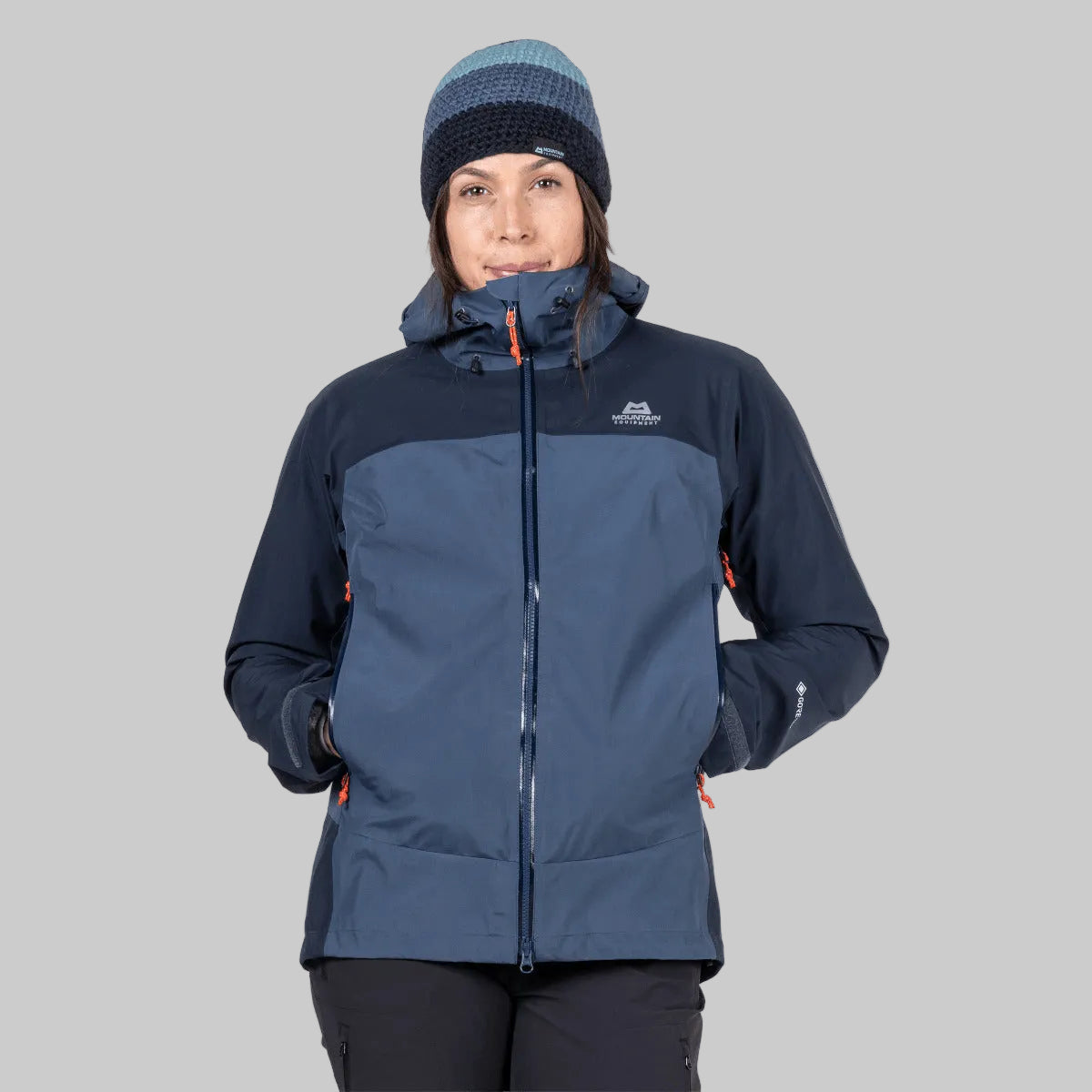 A woman wearing a dark blue and slate blue technical outdoor jacket with a matching striped beanie, photographed against a solid white background.