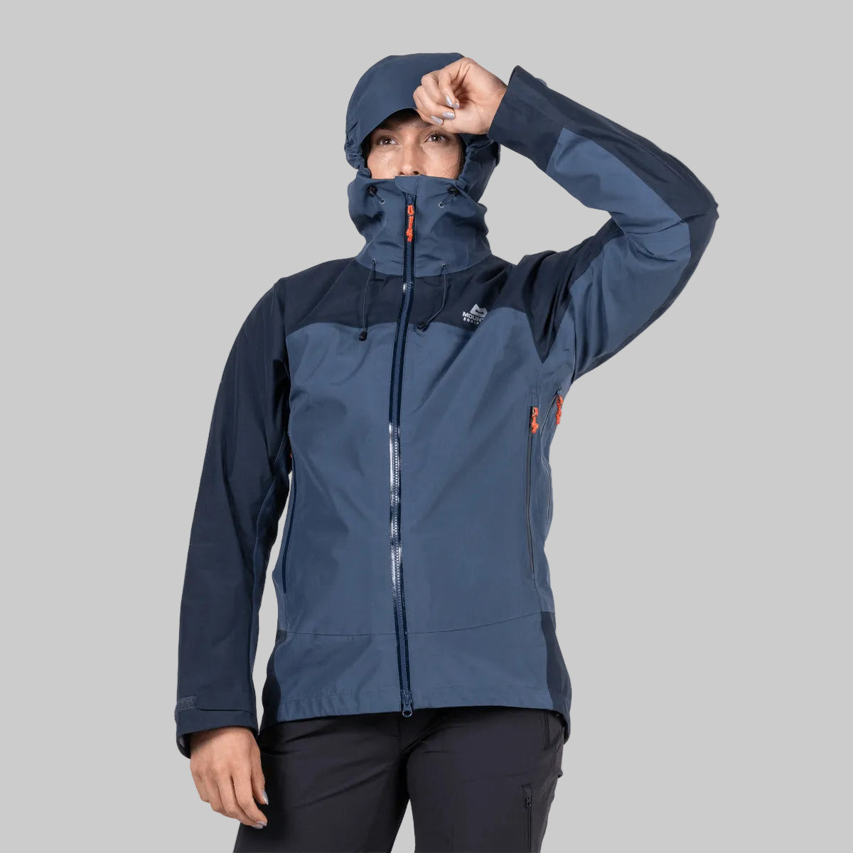 A woman wearing a navy and slate blue technical outdoor jacket with a high hood, posing against a solid white background.