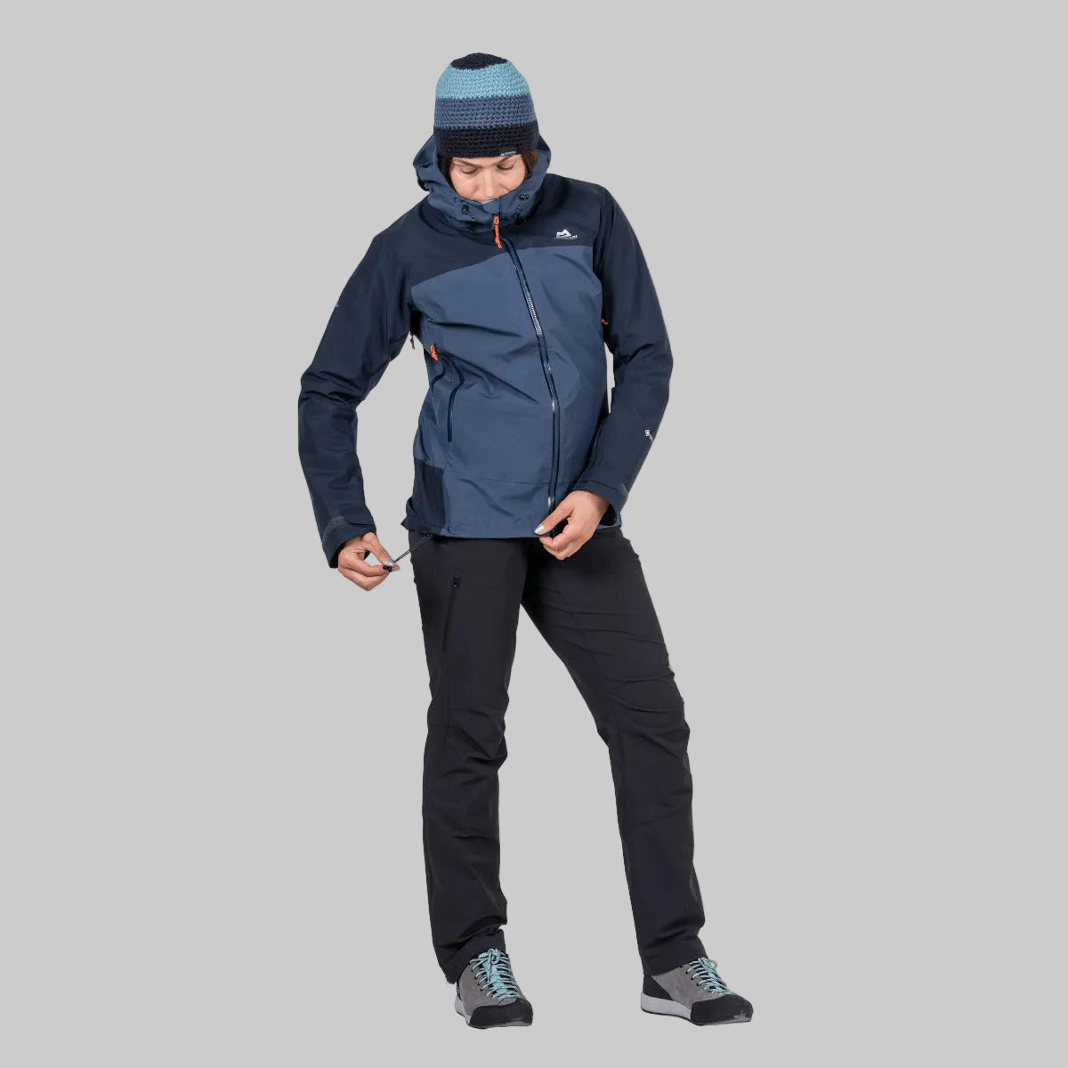 A man wearing a dark blue and navy outdoor jacket, a blue and gray striped beanie, black hiking pants, and gray hiking boots, standing against a solid white background.