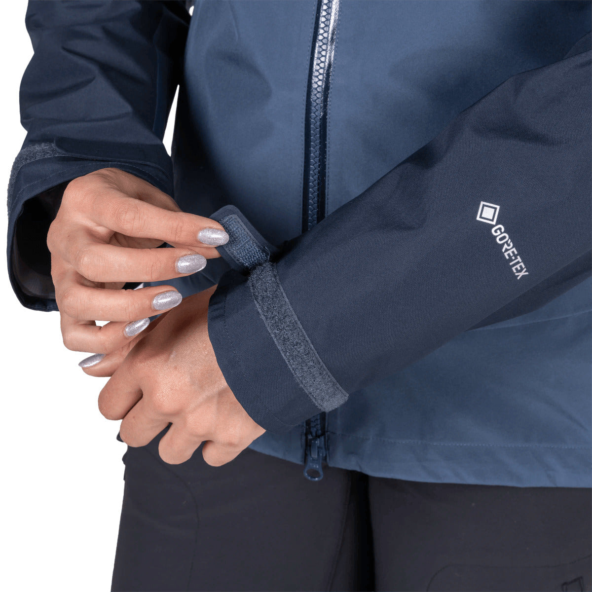 Close-up of a person wearing a dark navy Gore-Tex jacket, adjusting the sleeve cuff with both hands. The white Gore-Tex logo is visible on the upper left sleeve.