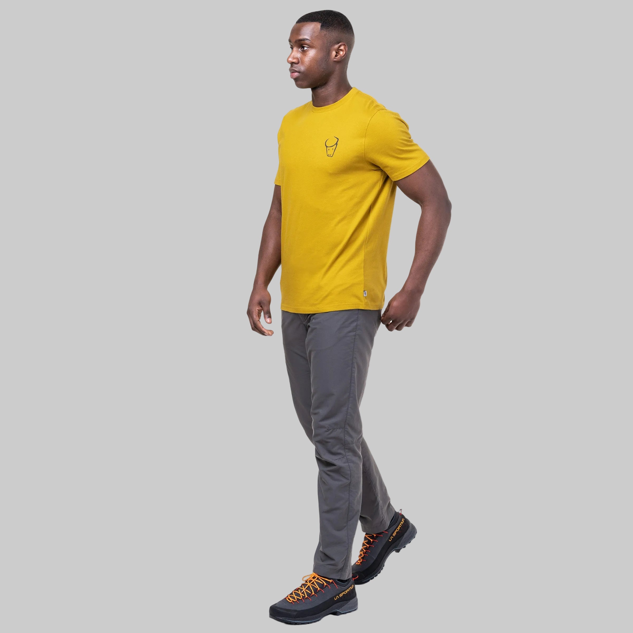 A man wearing a yellow short-sleeve athletic t-shirt with a small chest logo, gray pants, and black hiking shoes with orange laces, shown in profile against a solid white background.