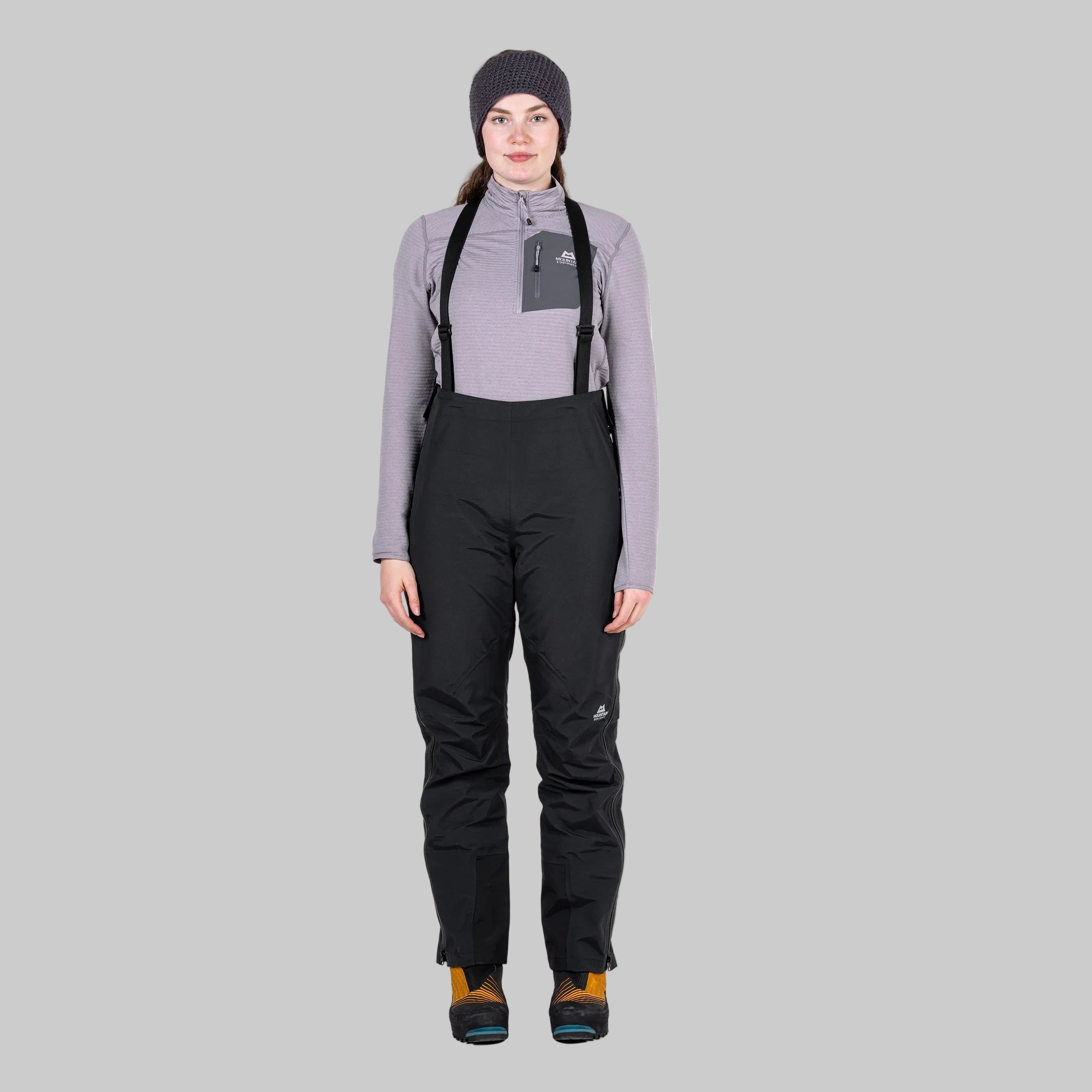 A woman modeling black ski bib pants with adjustable black suspenders, a light gray long-sleeve top, a dark gray beanie, and ski boots, photographed against a solid white background.