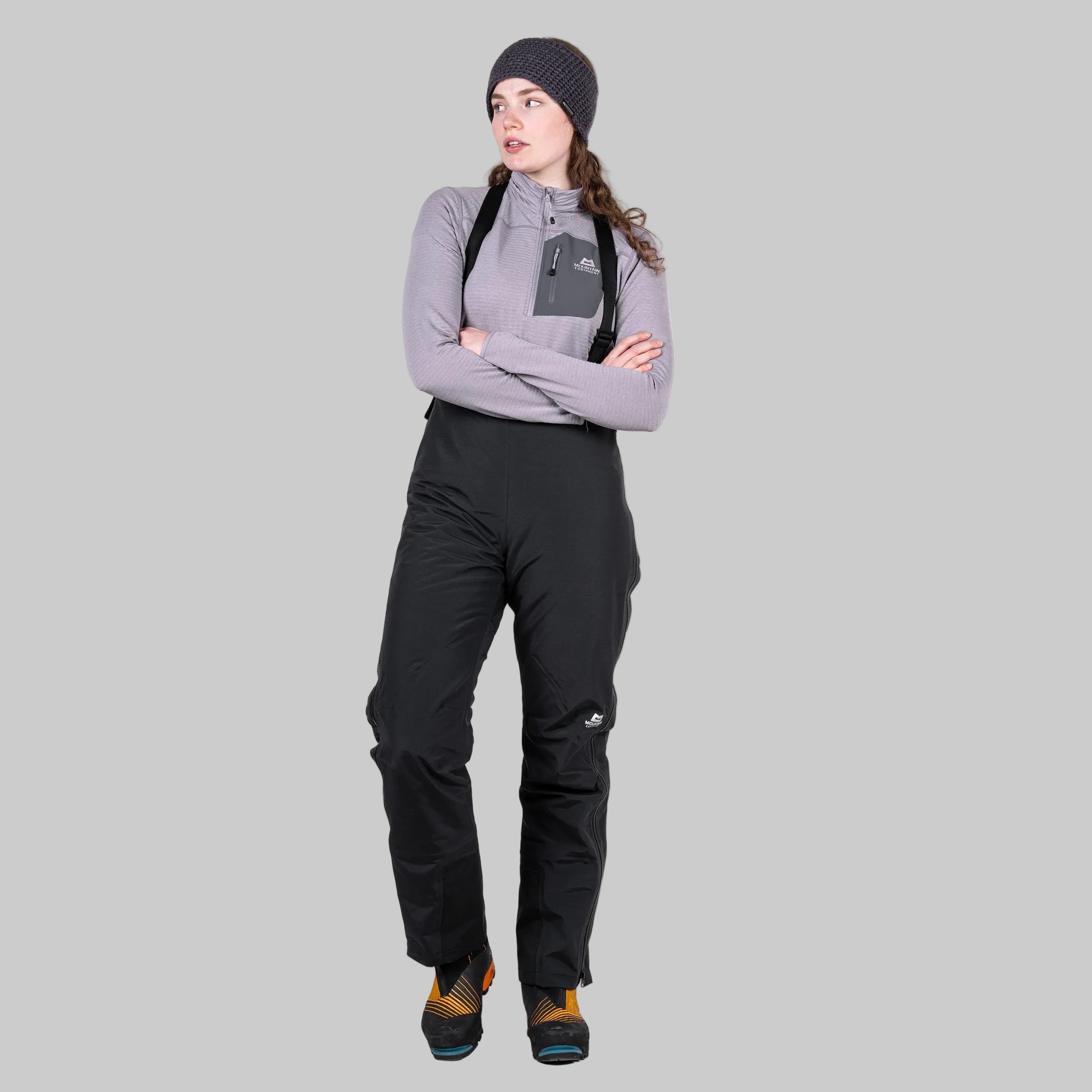 A woman modeling black ski bib pants with a gray long-sleeve top, black beanie, and hiking boots against a solid white background.