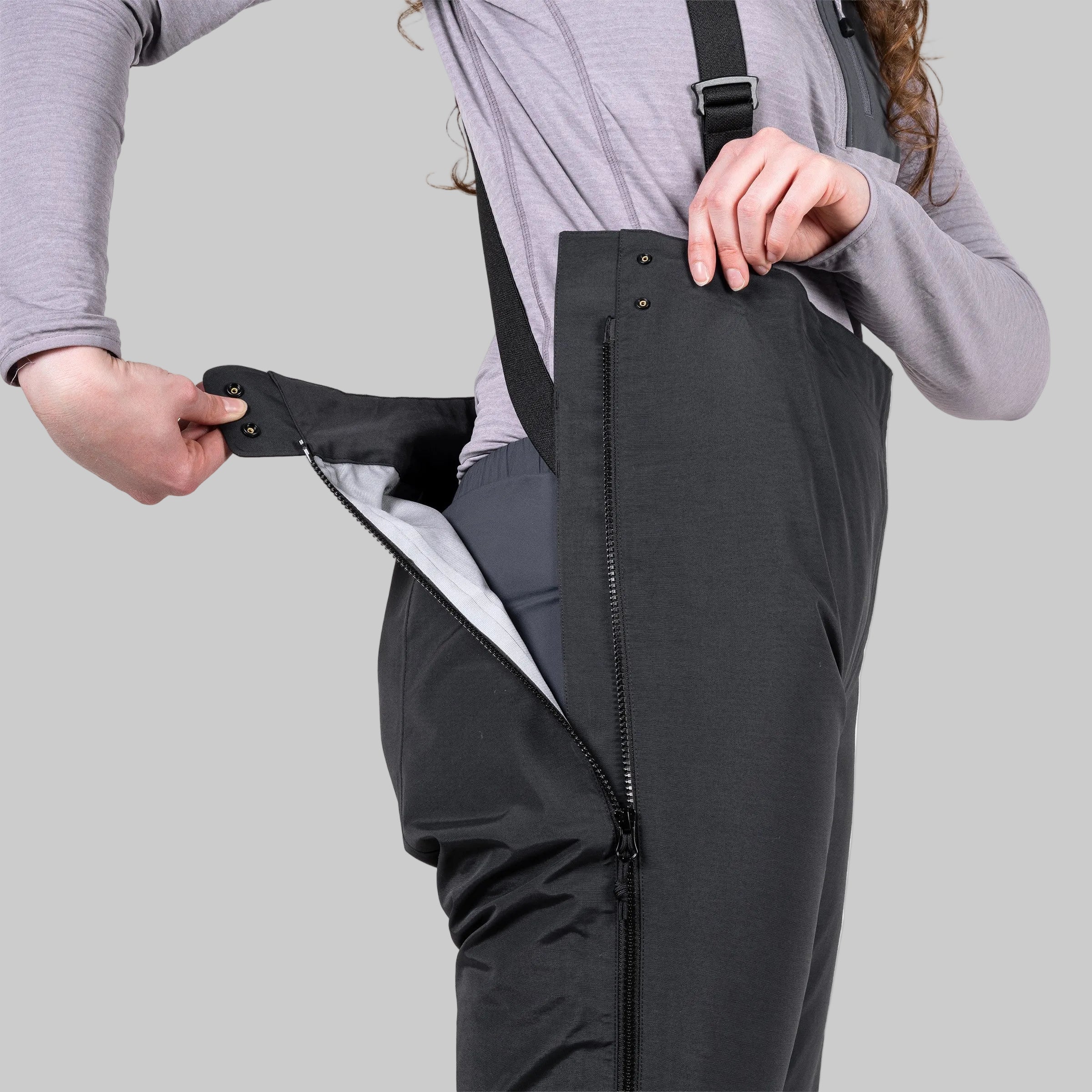 A person wearing black insulated bib overalls or snow pants, shown from the shoulders down, opening a large front zipper to reveal a light gray interior lining. The background is solid white.