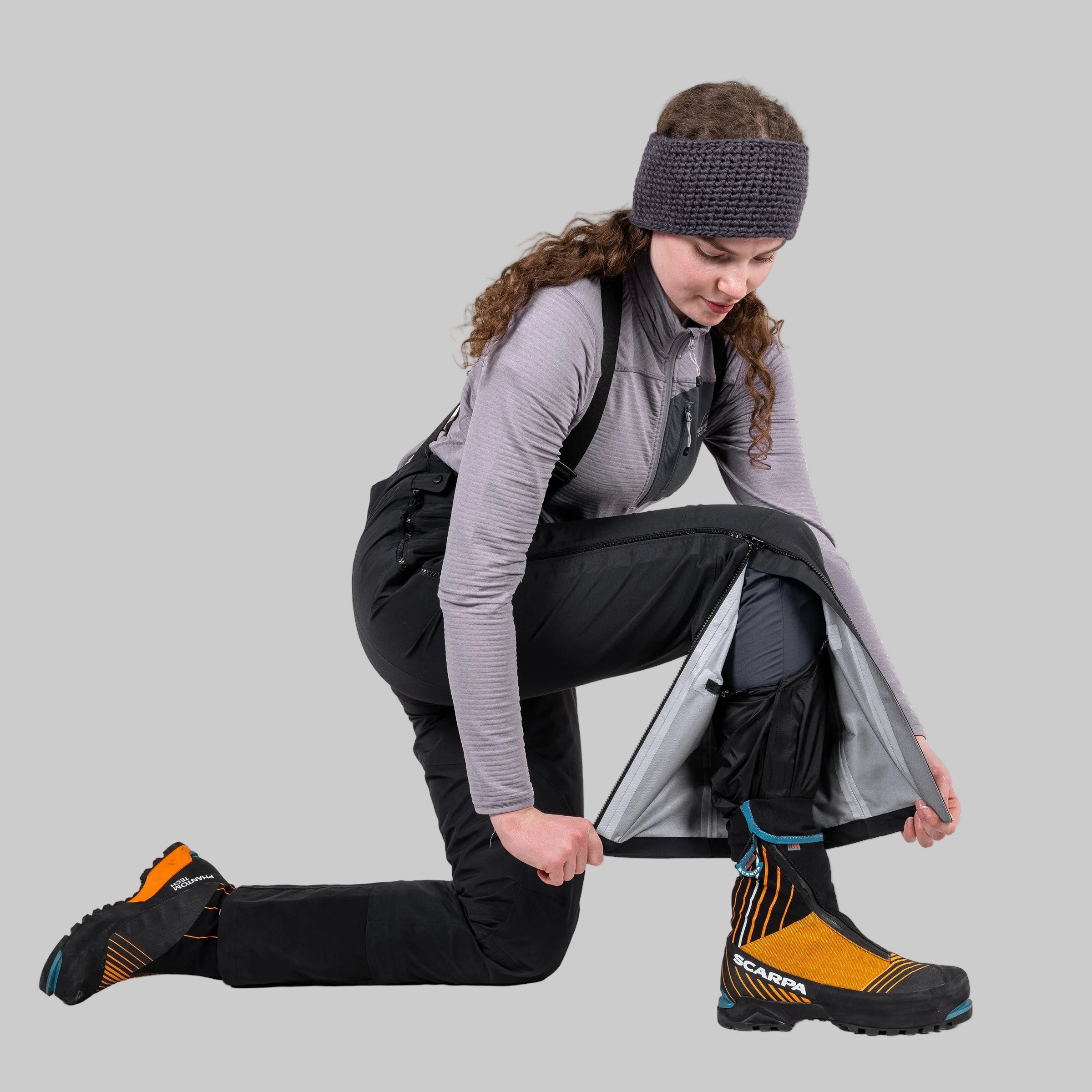 A woman kneels on a white studio background, demonstrating how to put on a pair of black insulated winter pants by pulling up a silver reflective cuff. She is wearing a gray long-sleeve top, a dark knit headband, and orange and black boot covers.
