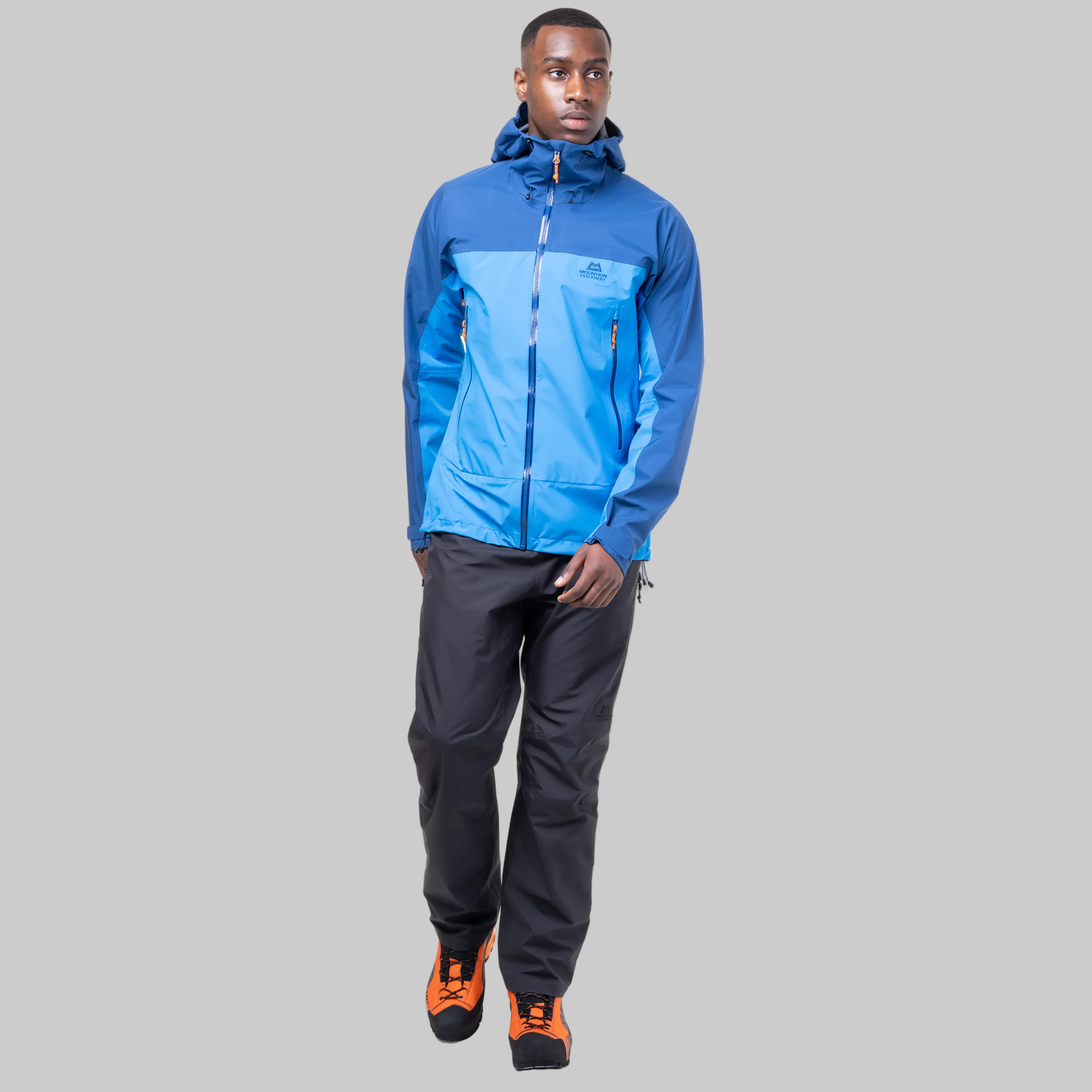 A man modeling a blue and light blue technical outdoor jacket with black pants and orange-and-black hiking boots, shown against a plain white background.