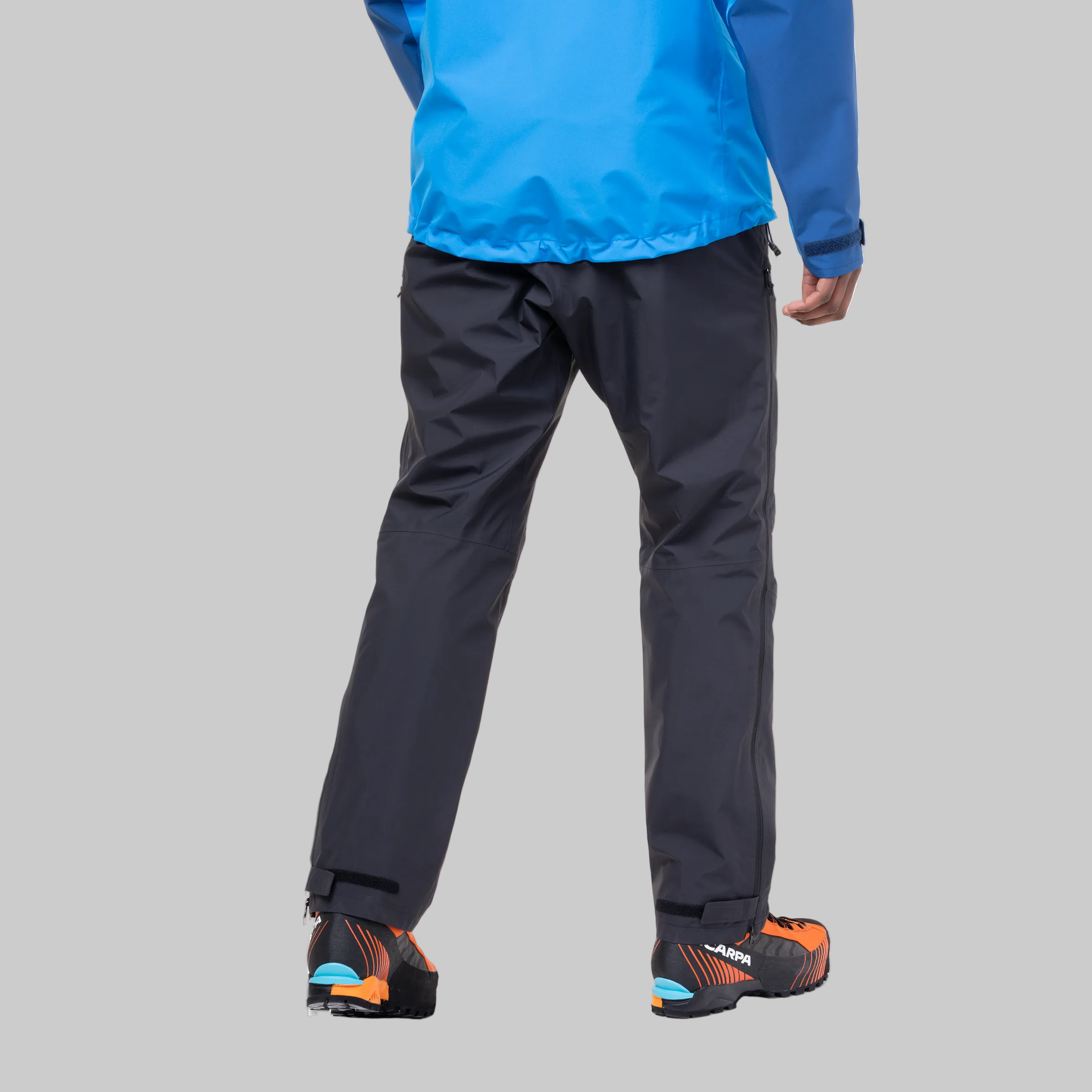 Rear view of a person wearing dark gray waterproof pants, a bright blue jacket, and orange and black ski boots against a grey background.