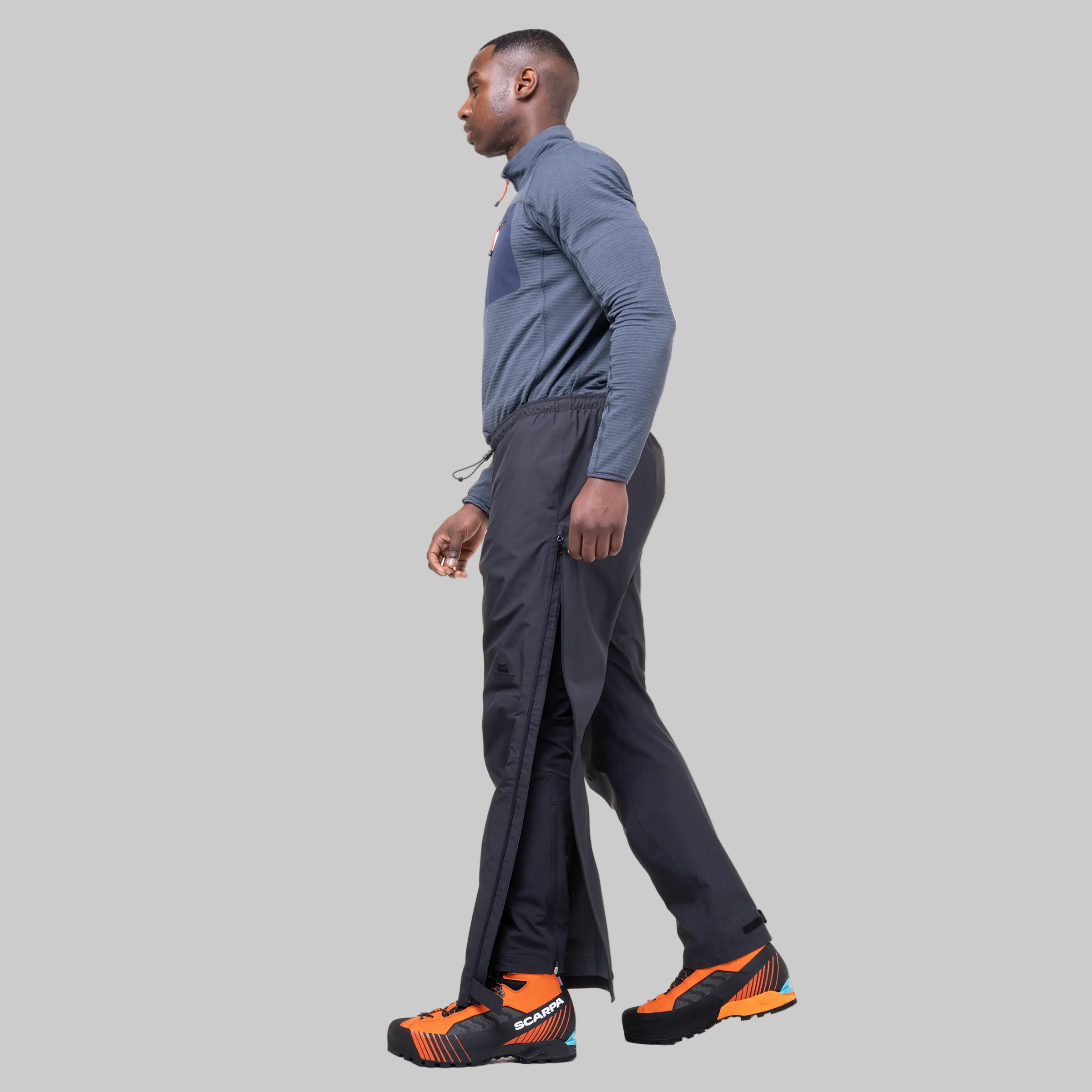 A man shown in profile wearing a slate blue long-sleeve athletic shirt, dark charcoal side-slit track pants, and orange and black trail running shoes against a plain white background.