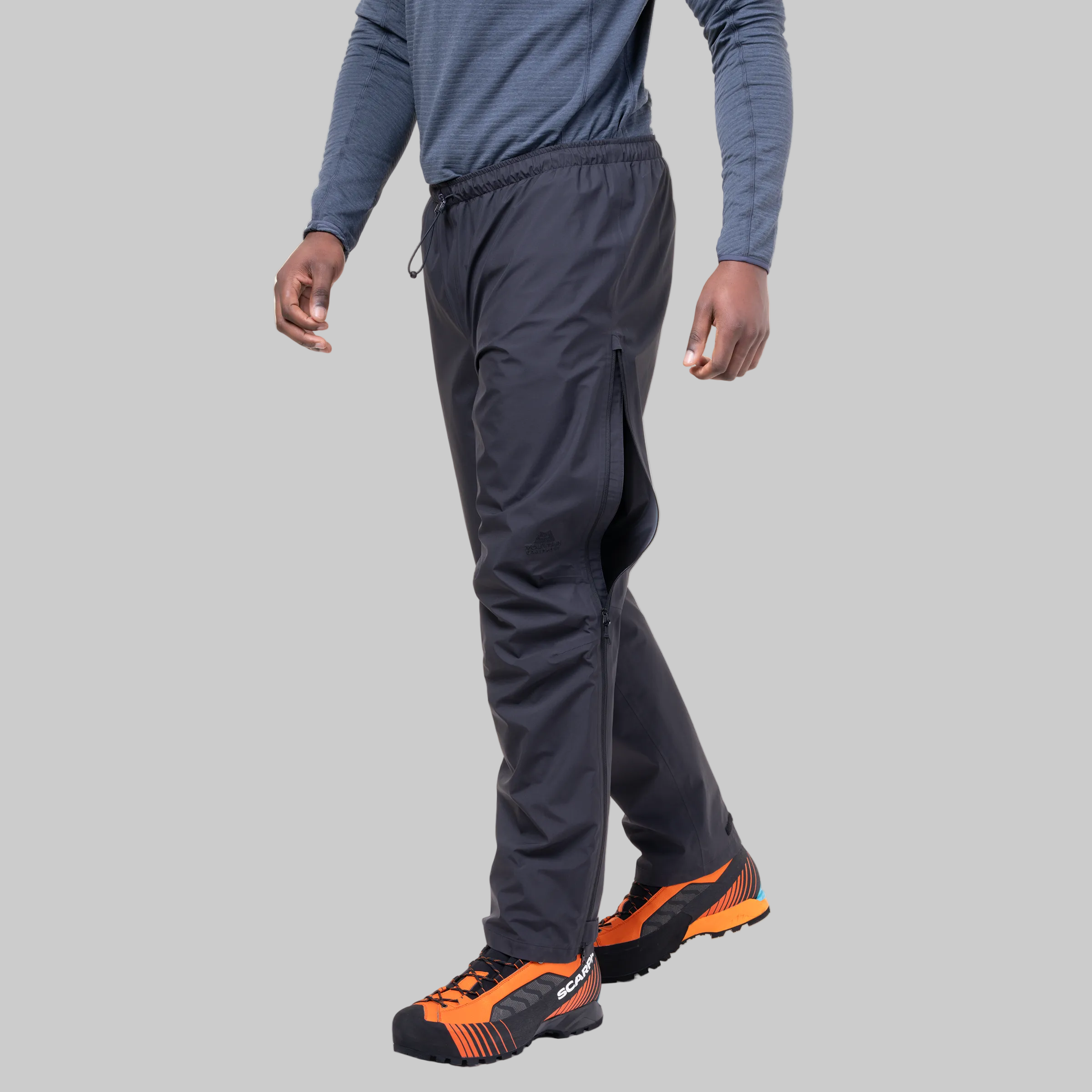 A person wearing dark charcoal zip-front athletic pants with a drawstring waist, paired with a blue long-sleeve shirt and orange-and-black trail running shoes, shown against a plain grey studio background.