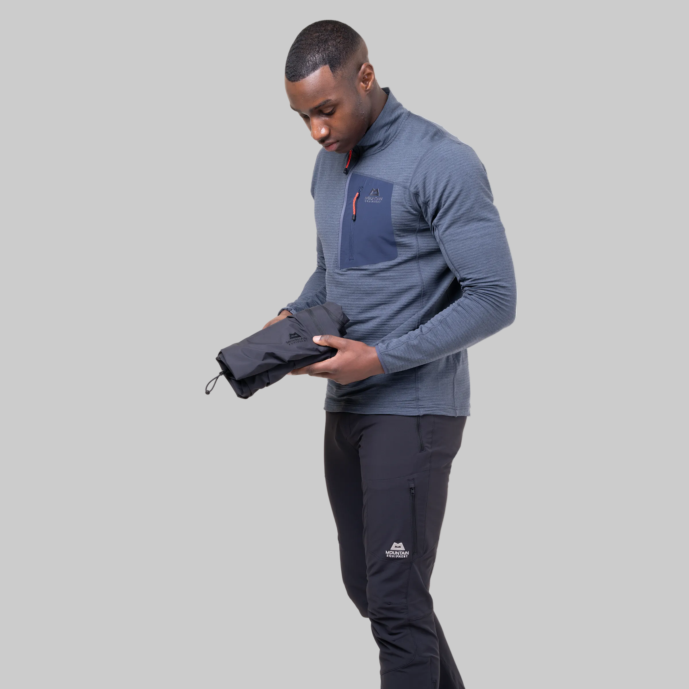 A man wearing a heathered slate blue quarter-zip pullover and black pants holds up a black drawstring pouch containing a matching dark gray pullover jacket. The background is solid white.