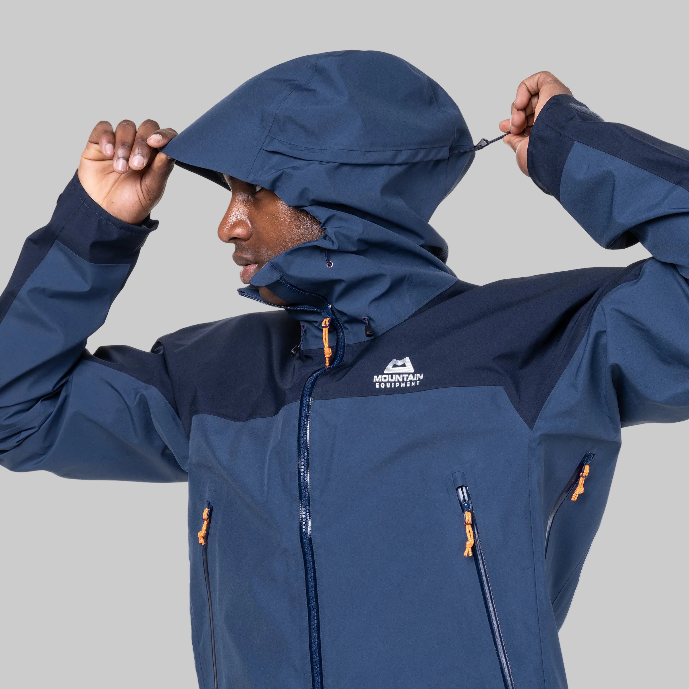 A man wearing a navy blue and dark navy waterproof jacket with orange zipper pulls, adjusting the hood. The jacket features a white 'MOUNTAIN EQUIPMENT' logo on the left chest.