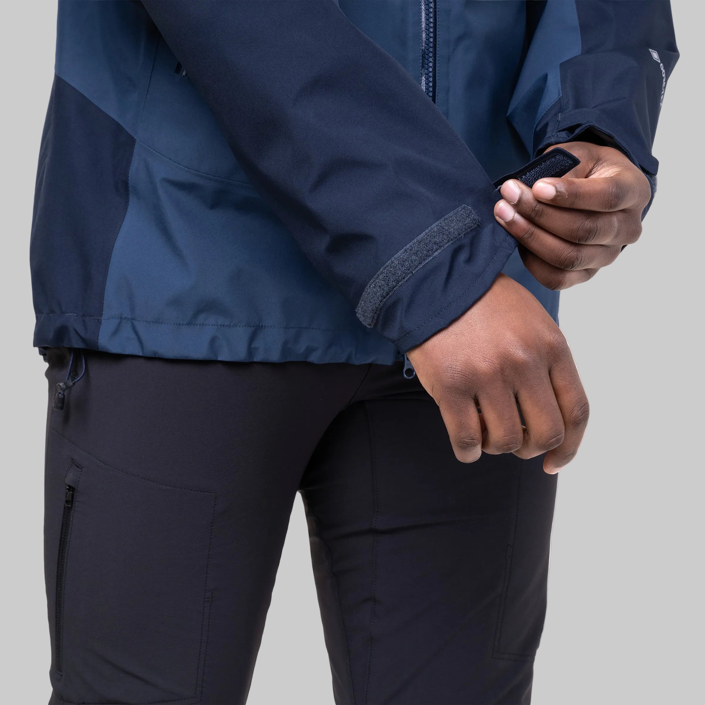 Close-up of a person wearing a navy blue technical jacket and dark pants, adjusting the cuff of the jacket with both hands against a clean white background.