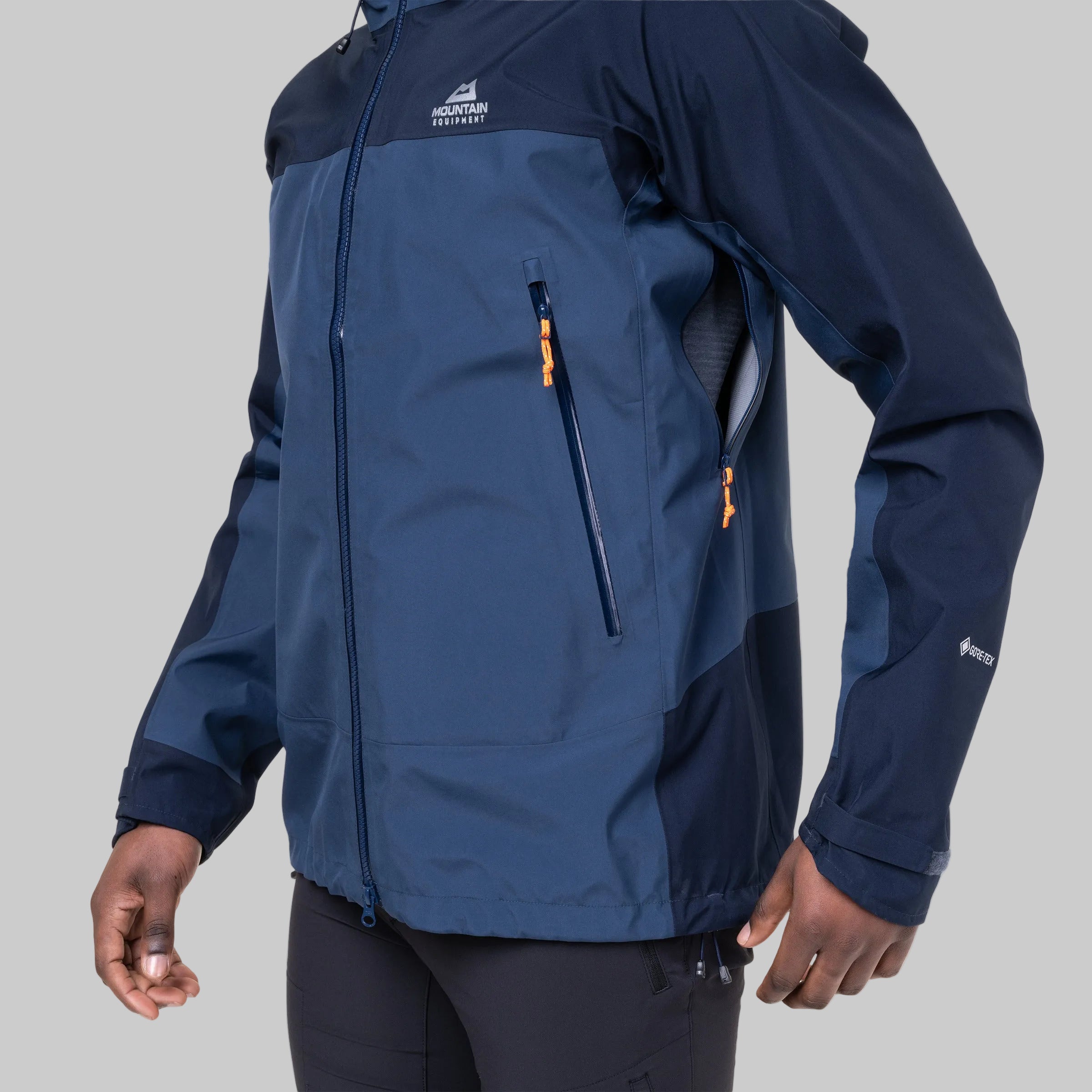 A man wearing a dark blue and navy technical outdoor jacket with orange zipper pulls, shown from the chest down against a white background.