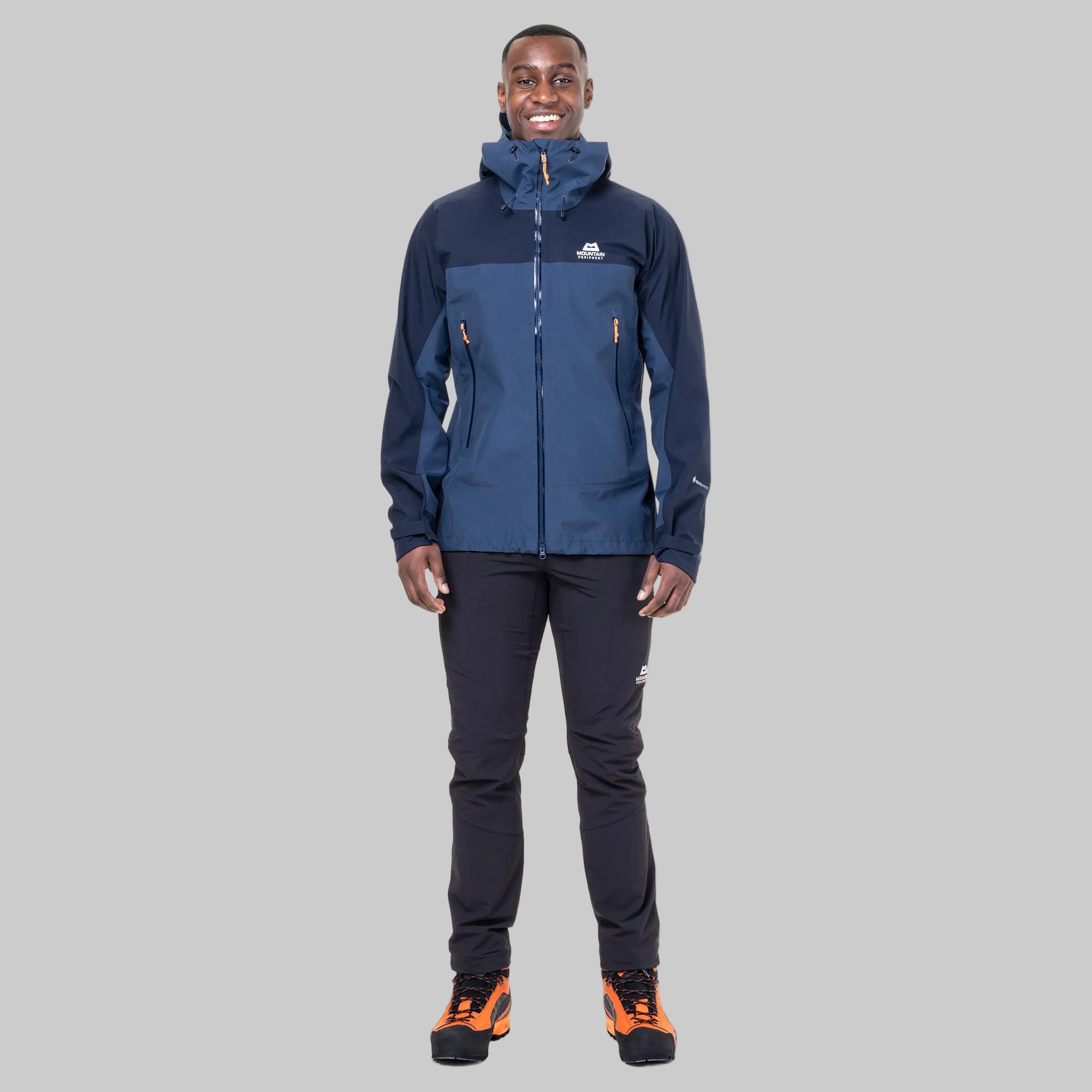 A man modeling a navy blue and dark blue technical outdoor jacket with orange zipper accents, paired with black pants and orange/black hiking boots, against a solid white background.