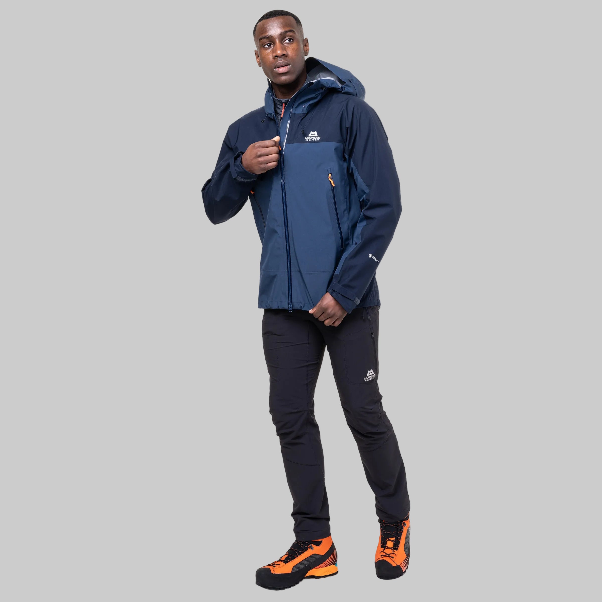 A man modeling a navy blue hooded technical jacket with a blue front panel, paired with black pants and orange and black hiking boots, against a solid white background.