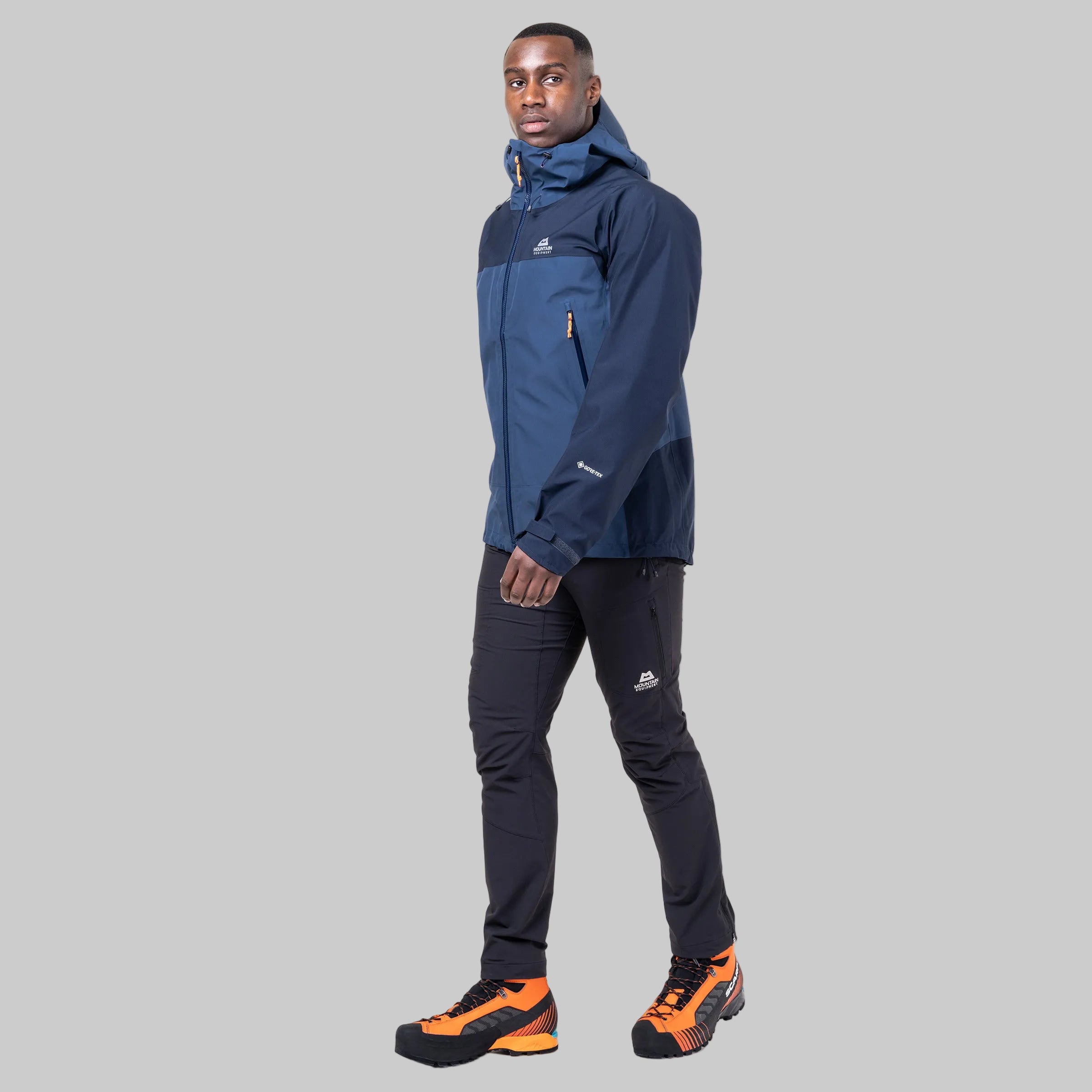 A man modeling a navy blue and dark blue technical jacket, paired with dark pants and orange and black hiking boots, against a plain white background.