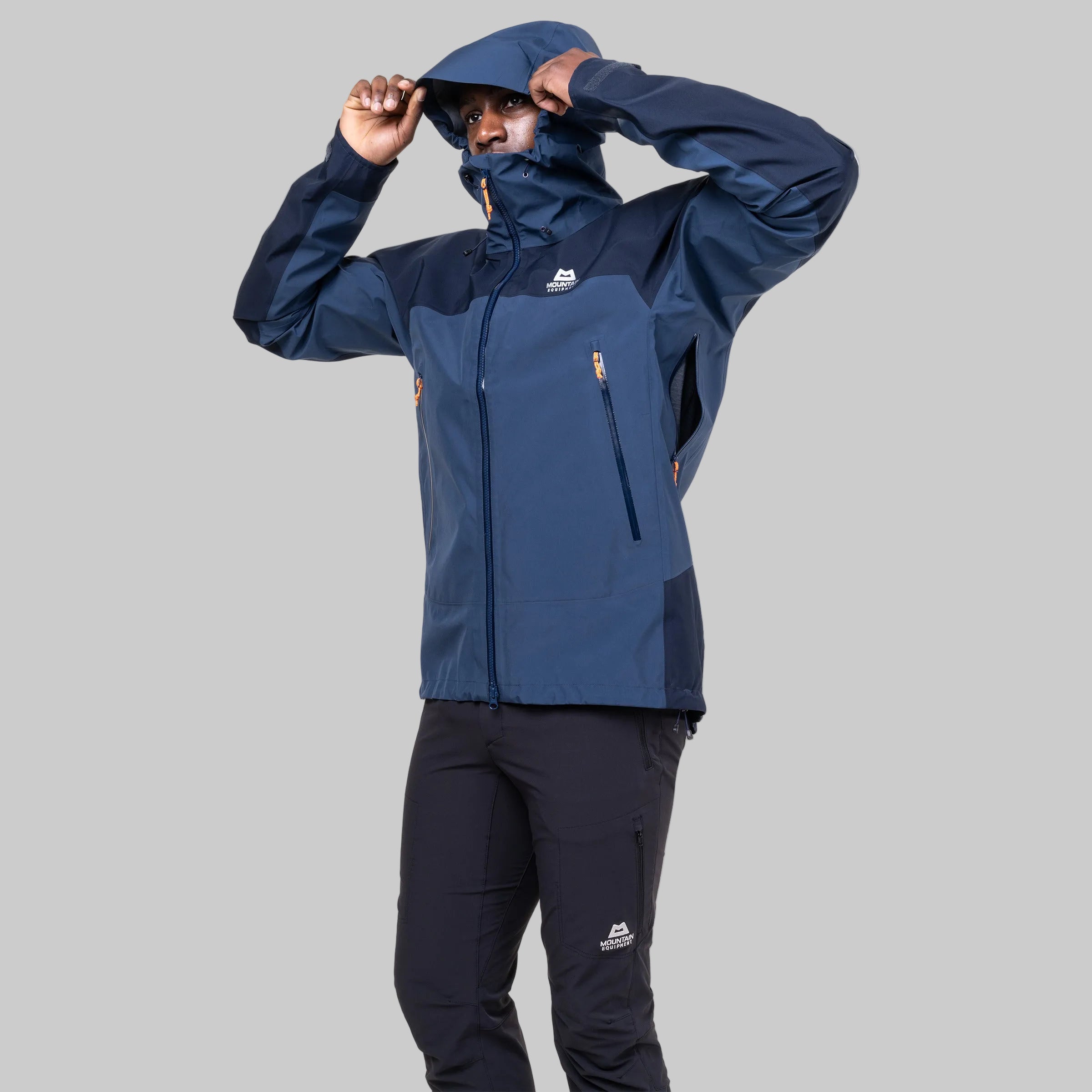 A man wearing a navy blue and dark blue waterproof jacket with a matching hood, holding the hood up over his head, paired with dark pants against a solid white background.