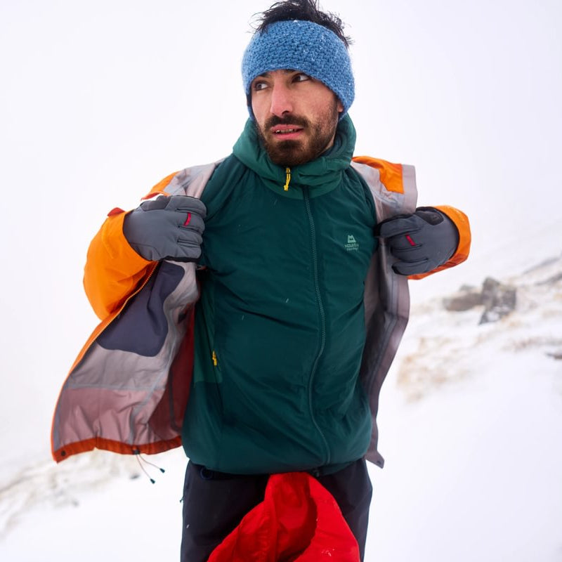 A man in a snowy outdoor setting wearing a dark green technical jacket, an orange and gray shell layer draped off his shoulders, a blue knit beanie, and gray gloves.