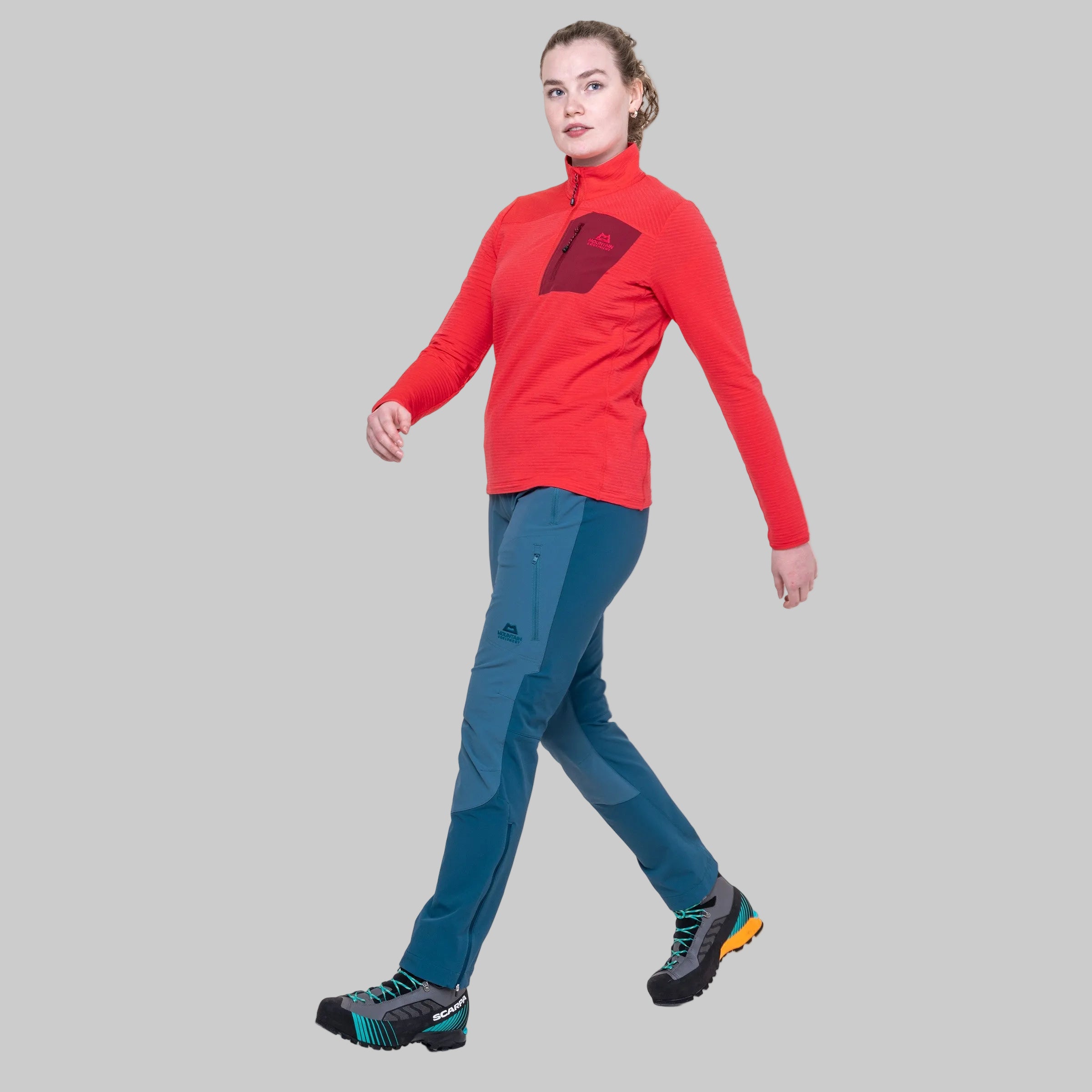 A woman modeling a red long-sleeve quarter-zip athletic top and teal hiking pants against a solid gray background.
