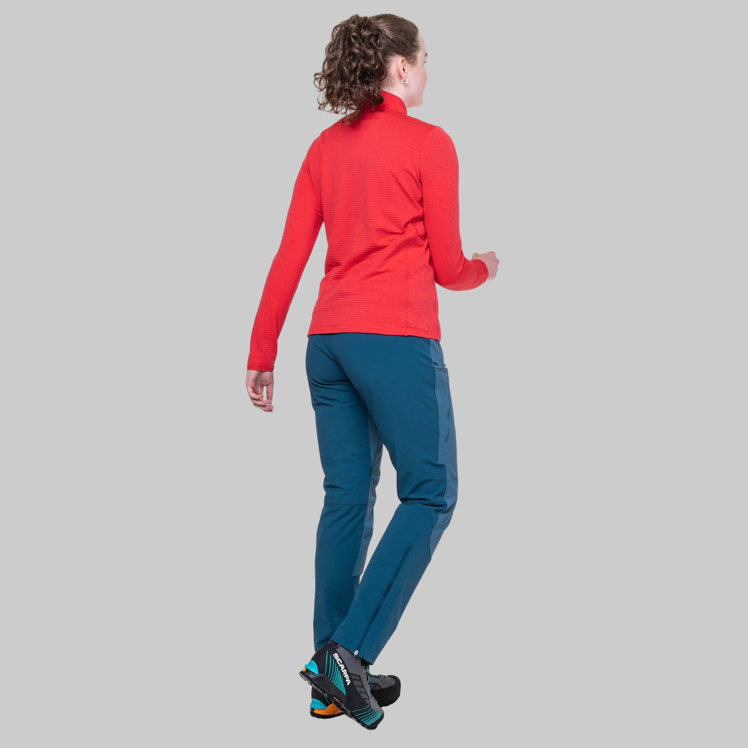 A woman shown from the back wearing a bright red long-sleeve athletic top and teal blue pants, standing against a plain gray background.