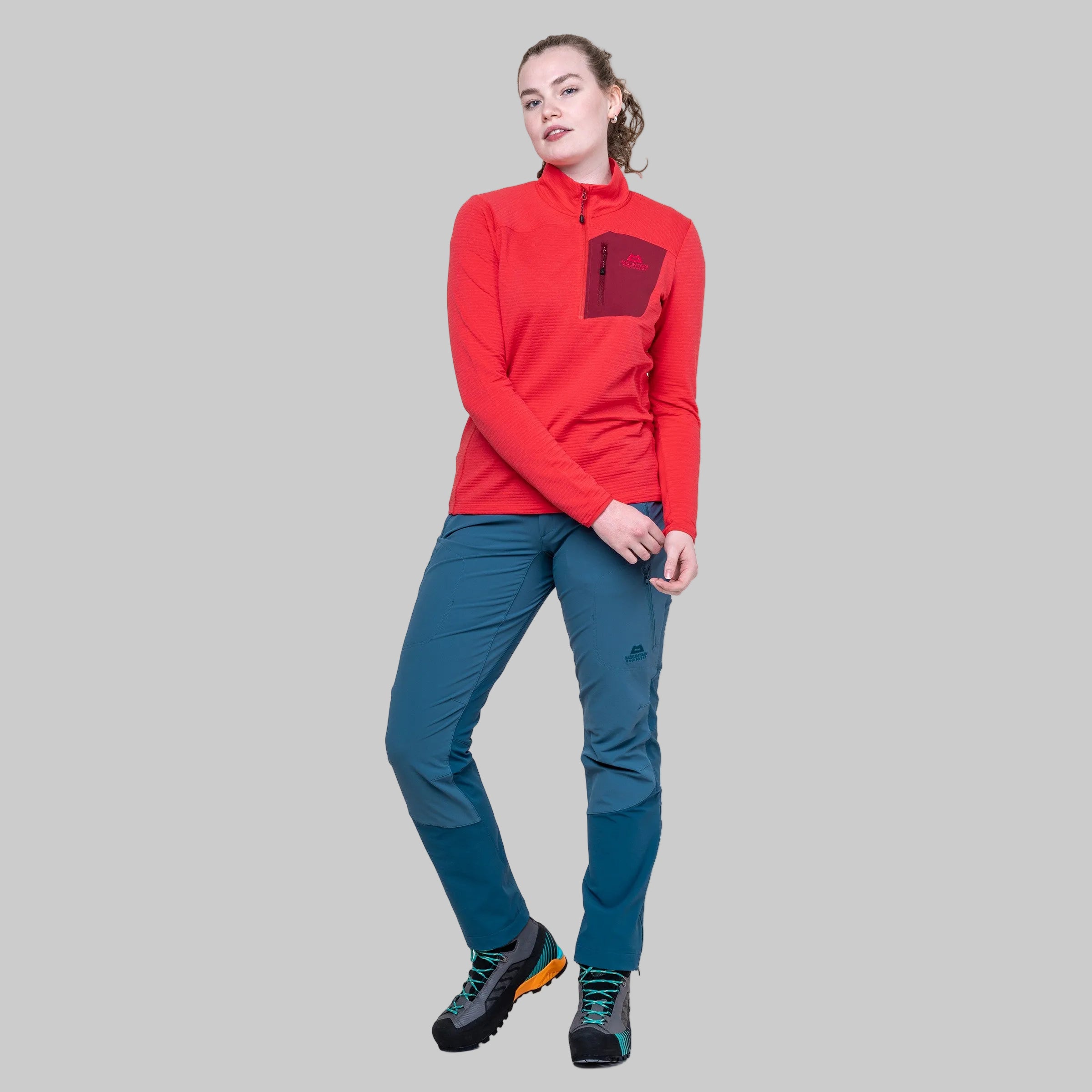 A woman modeling a red long-sleeve quarter-zip fleece top and teal hiking pants against a solid white background.