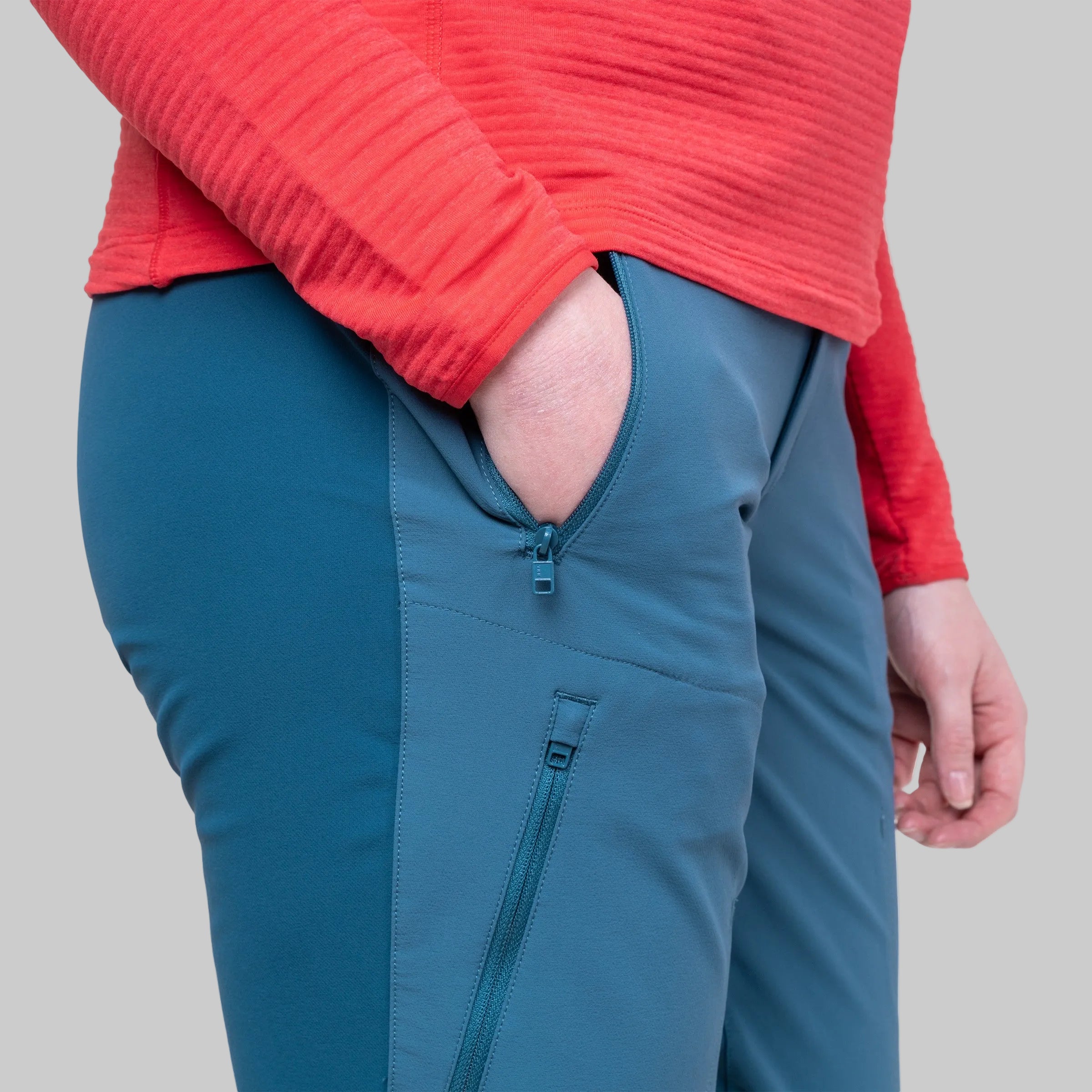 Close-up of a person wearing teal blue technical pants with a zippered side pocket and a red ribbed long-sleeve top, shown against a plain gray background.