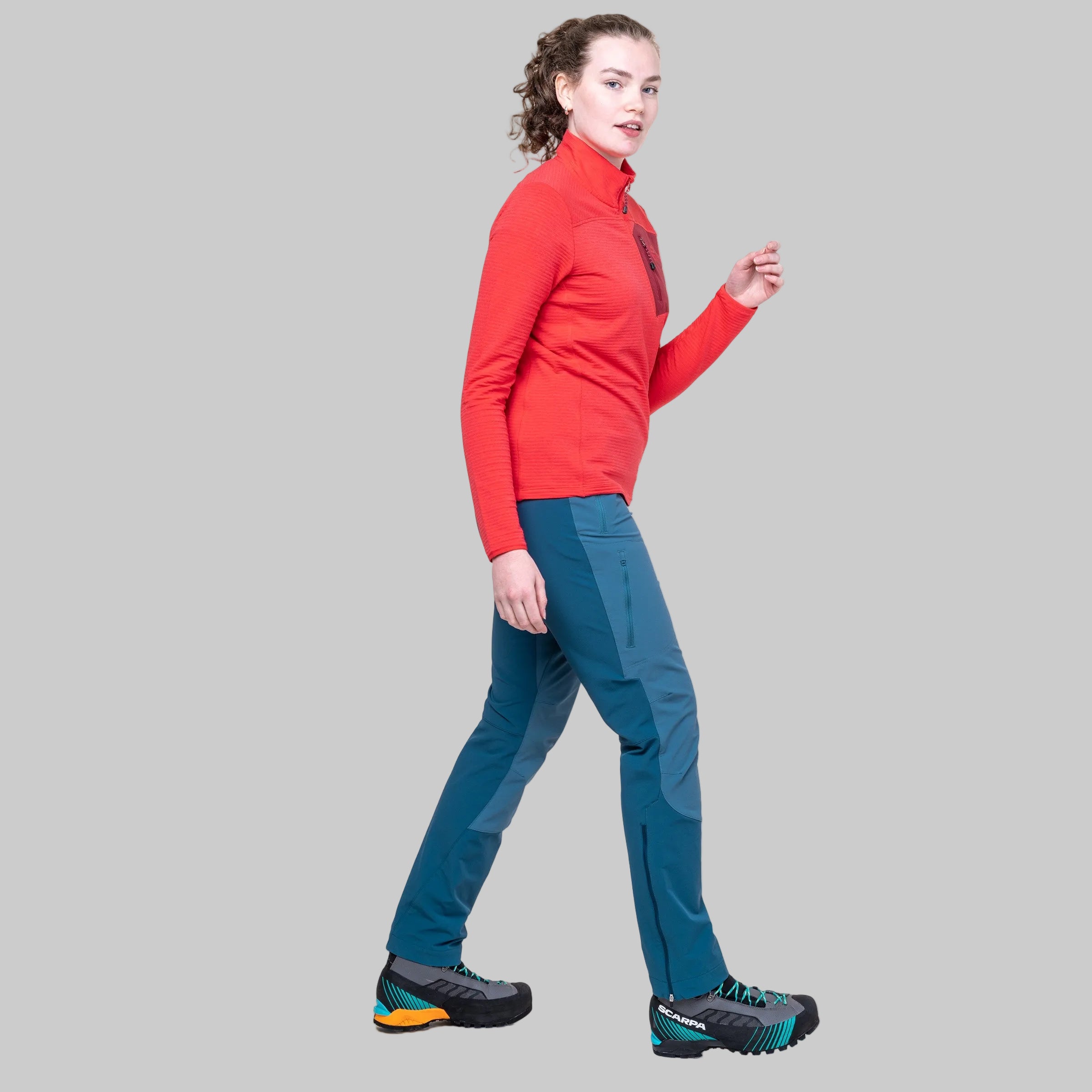 A woman modeling a red long-sleeve quarter-zip athletic top and teal hiking pants with black and teal trail shoes, shown in profile against a solid gray background.