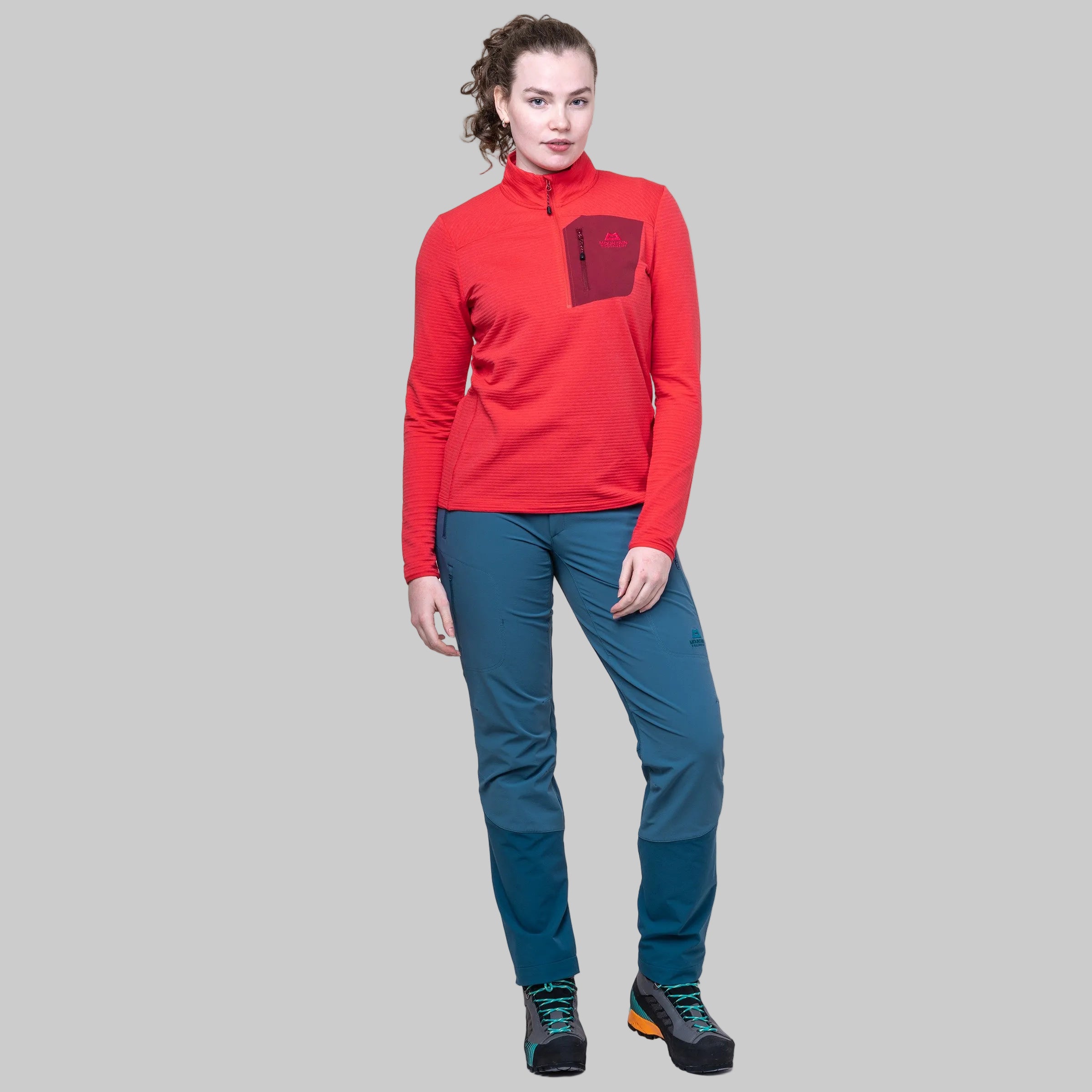 A woman modeling a red long-sleeve quarter-zip fleece with a chest pocket, paired with teal hiking pants and black hiking boots, against a solid white background.