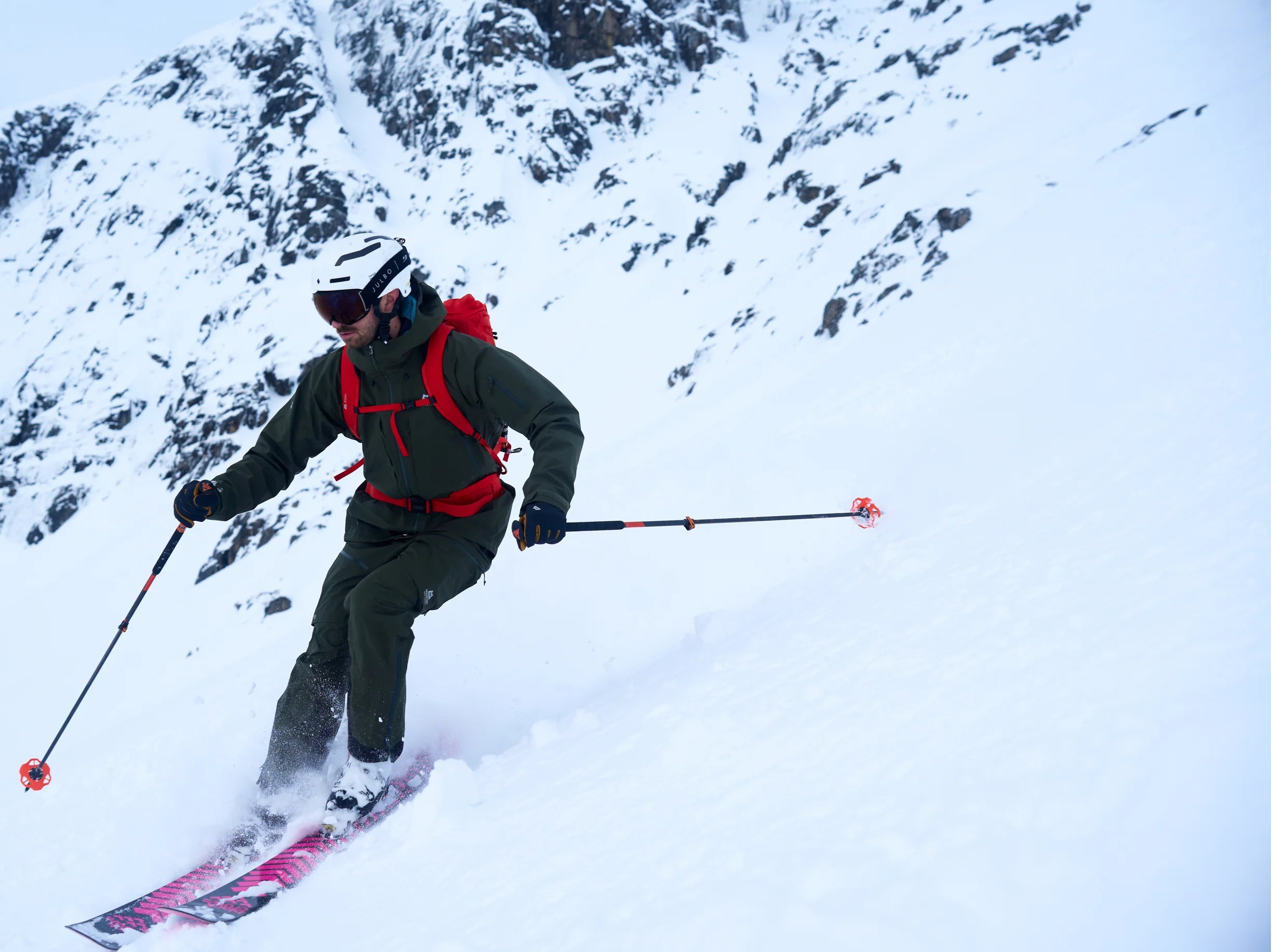 A skier carving through deep snow on a steep mountain slope, wearing a dark green ski jacket and pants with a red backpack, helmet, and goggles, holding ski poles.