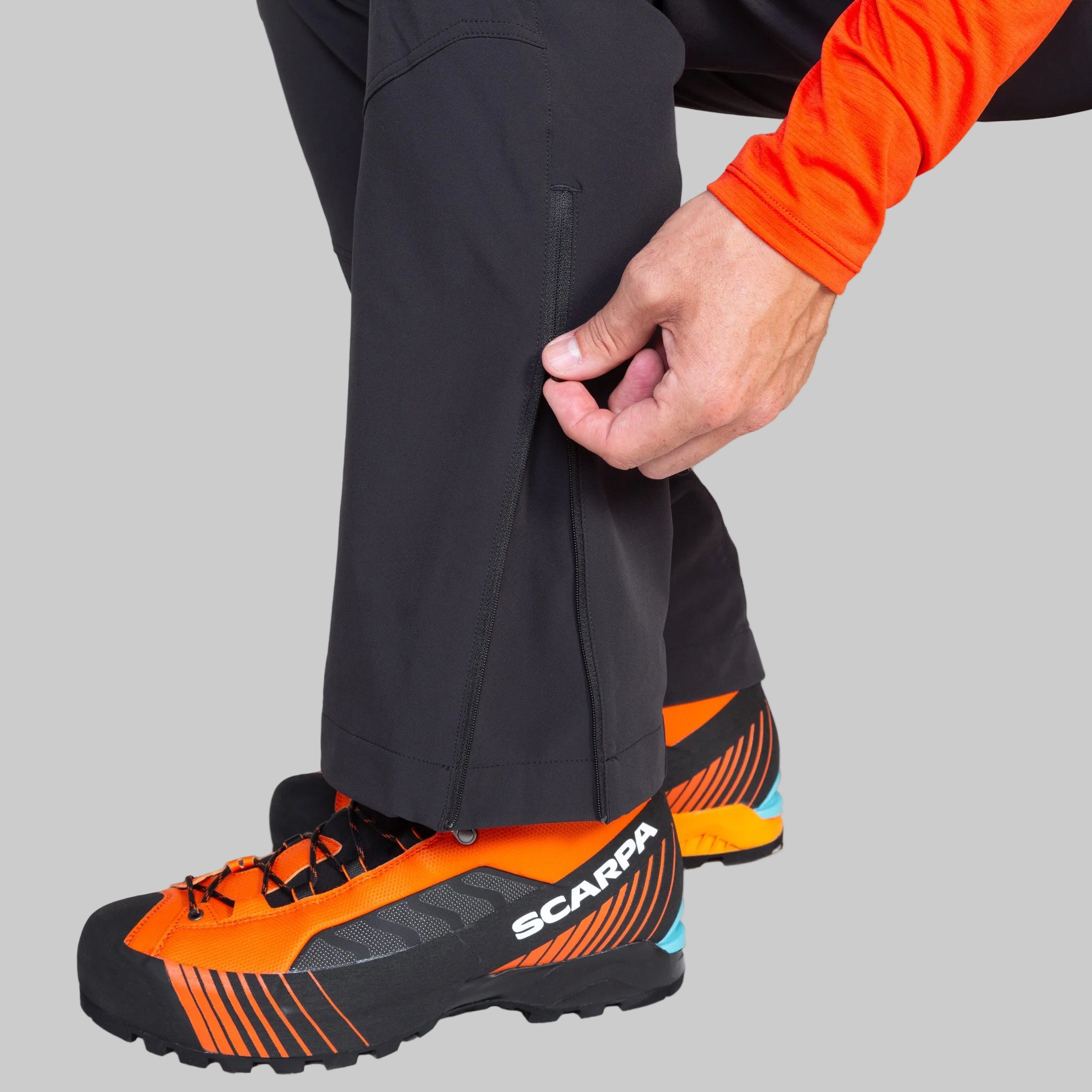 A close-up of a person's leg and hand adjusting black pants, with orange and black Scarpa hiking shoes visible against a light gray background.