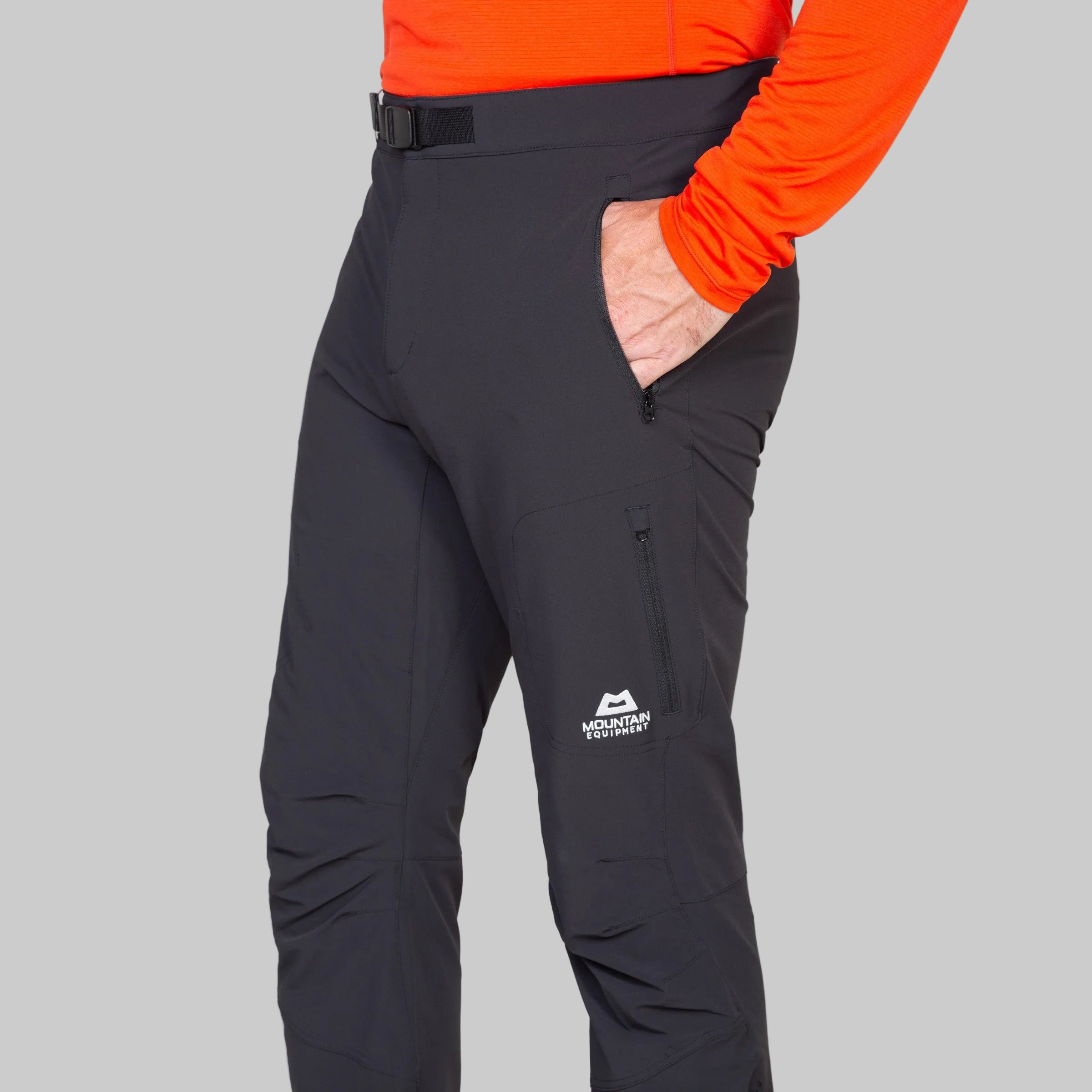 A close-up of a person wearing black technical pants with a white Mountain Technology logo on the lower left leg, paired with a bright orange long-sleeve top. The person's hand is in their pocket, set against a plain light gray background.