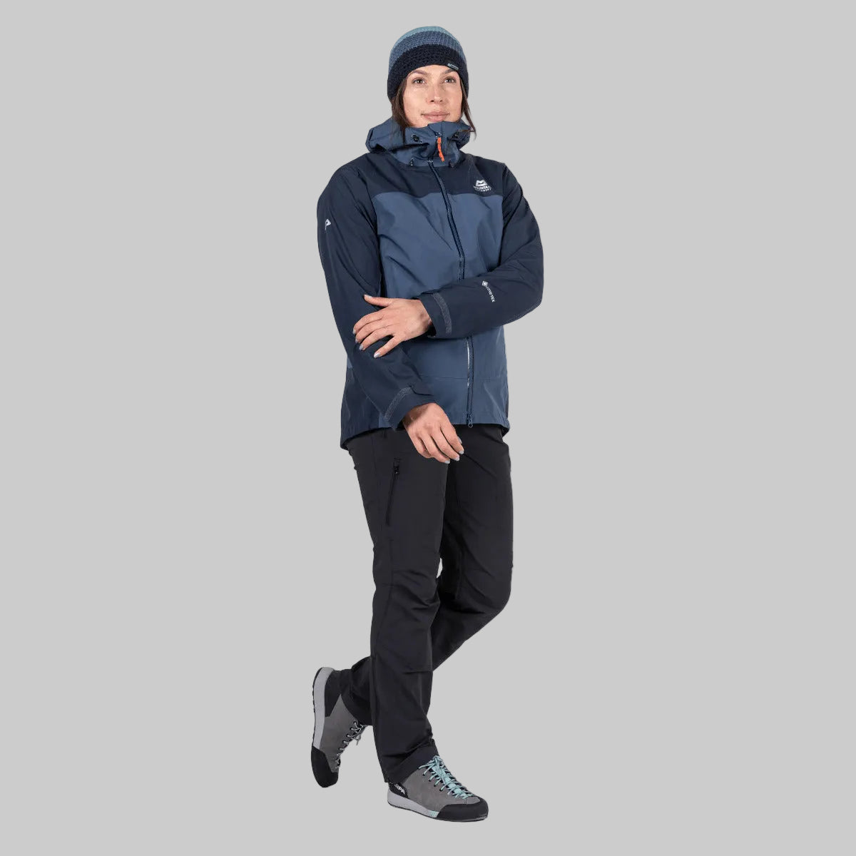 A man modeling a dark navy and slate blue technical outdoor jacket, paired with black pants, a dark beanie, and hiking boots, against a solid white background.