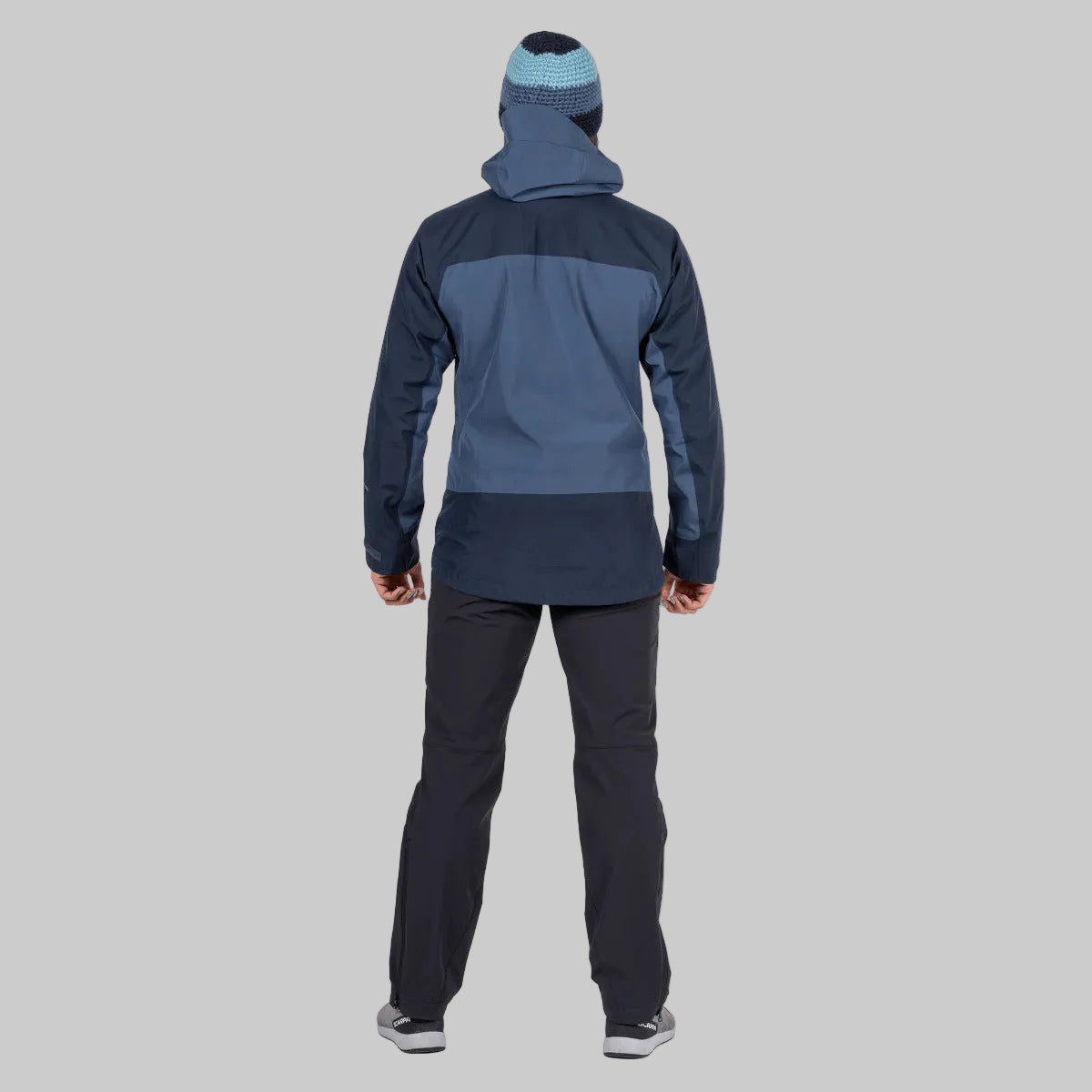 A person shown from the back wearing a colorblock hooded jacket in navy and slate blue, dark pants, and a blue and black beanie on a solid white background.