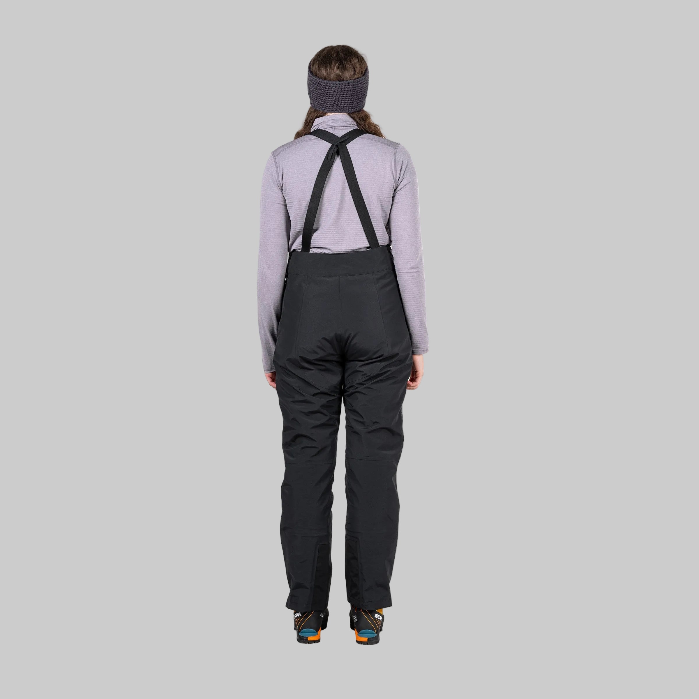 A person shown from the back wearing black bib overalls with black suspenders, a light gray long-sleeve shirt, a dark knit headband, and dark sneakers, against a plain white background.